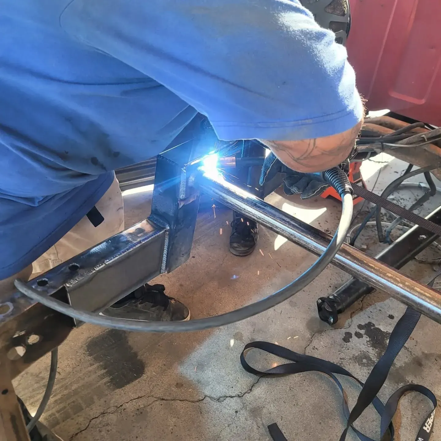 A person welds a metal tube to a vehicle frame, wearing a blue shirt and protective gear in an outdoor setting.