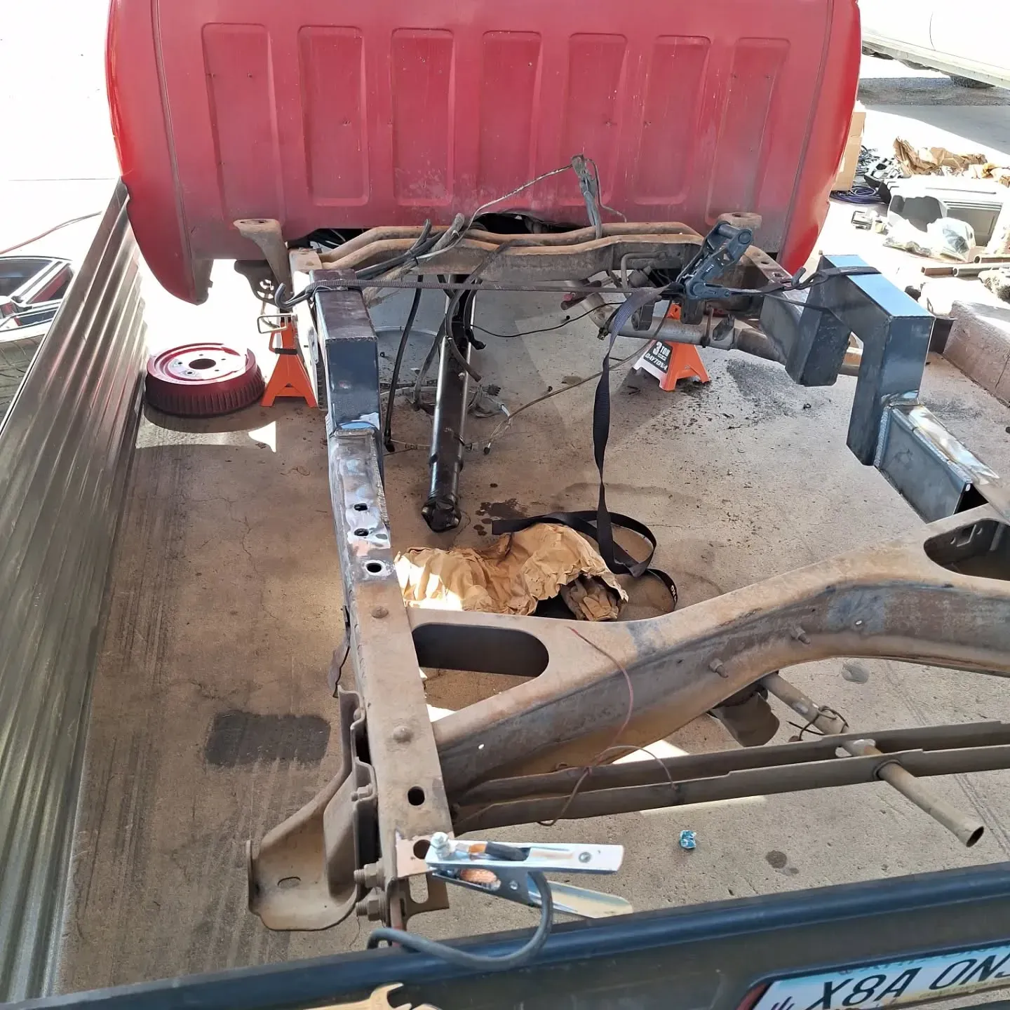 Red truck bed and exposed chassis, undergoing repair outdoors. A damaged section is visible, with tools and supports nearby.