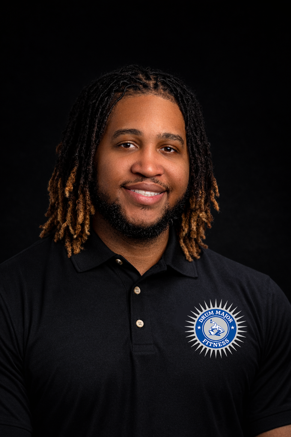 A person with shoulder-length dreadlocks smiles while wearing a black polo shirt featuring a circular white and blue emblem.