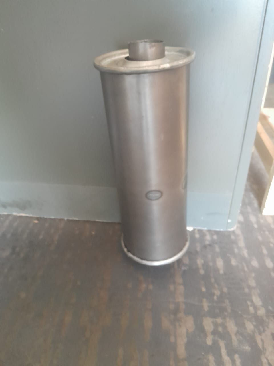 Silenciador de automóvil cilíndrico de metal sobre un piso gris contra una pared.