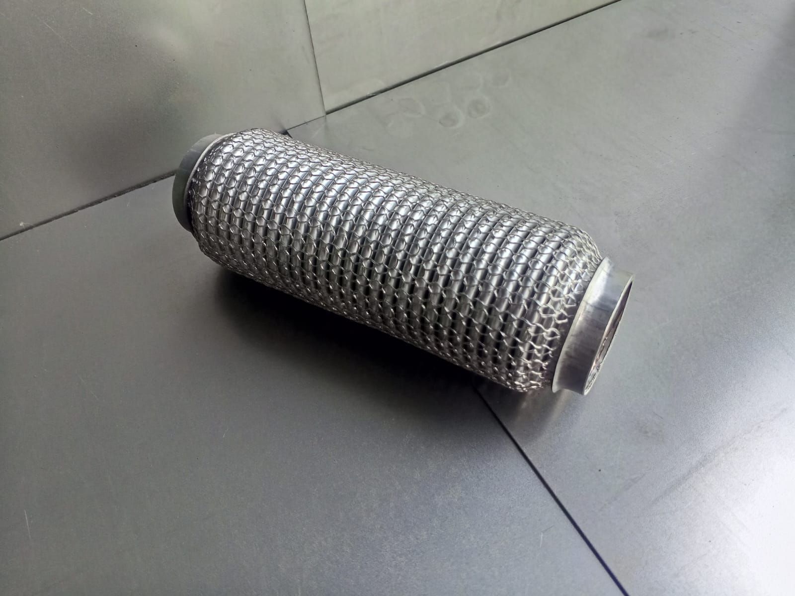 Tubo de escape flexible con exterior de metal tejido, sobre superficie plateada.