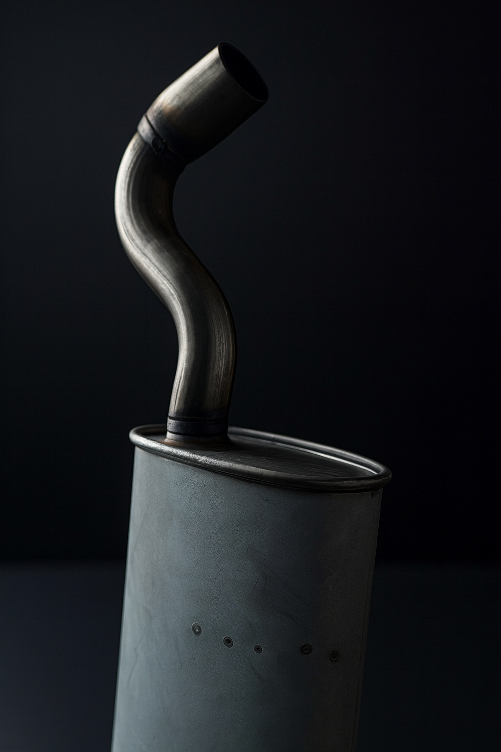 Un primer plano de un silenciador de automóvil gris con un tubo de escape curvo y negro sobre un fondo negro.