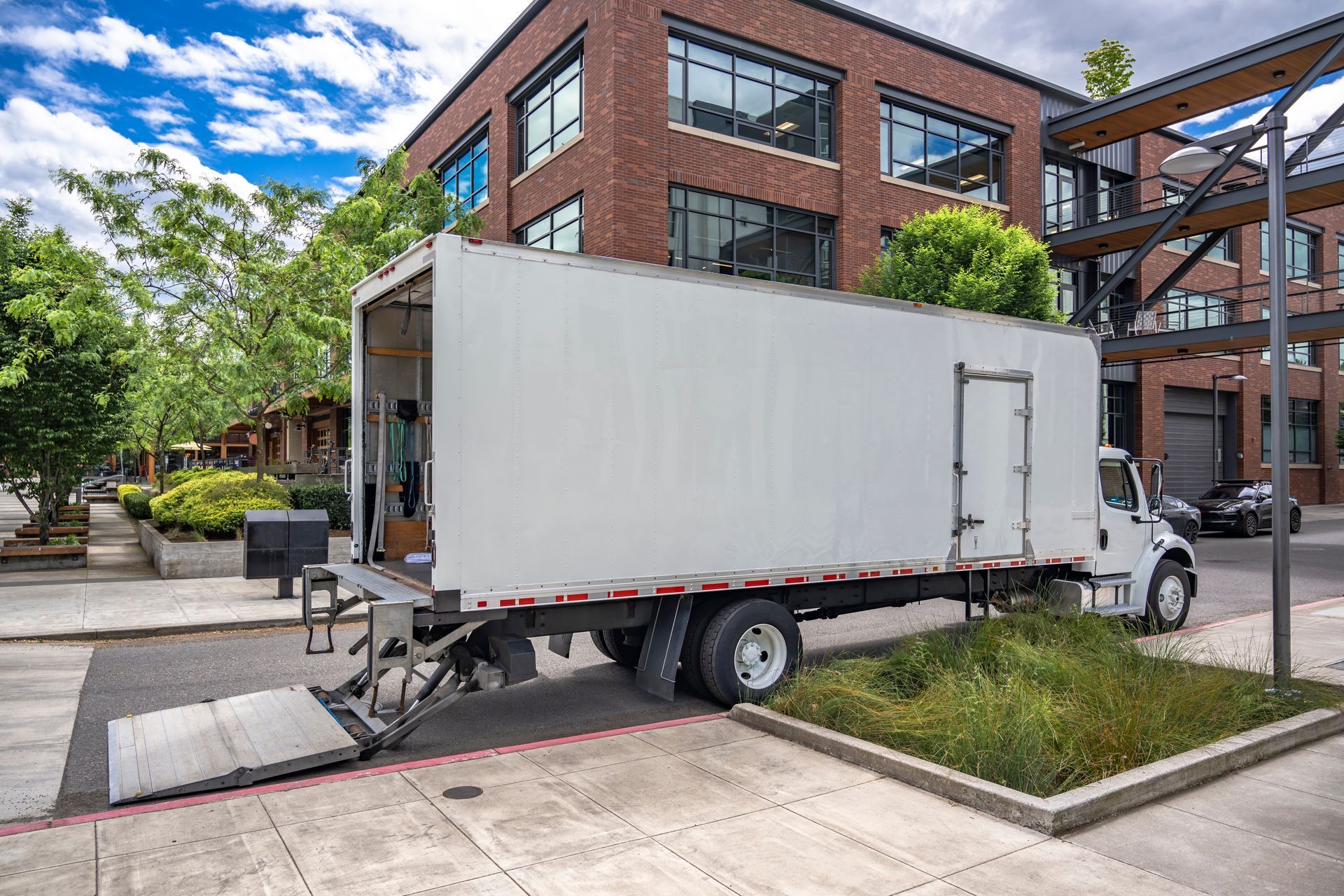 A white moving truck is parked on the side of the road in front of a brick building.