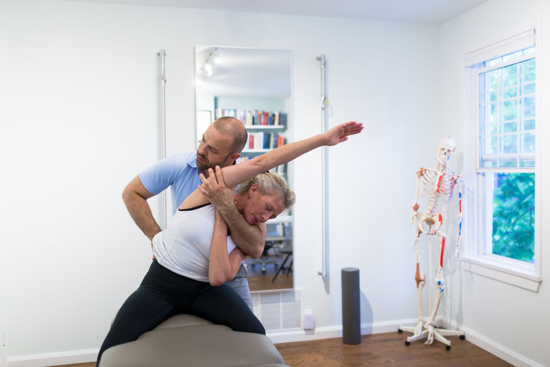 A man is helping a woman stretch her arms while sitting on a yoga ball.