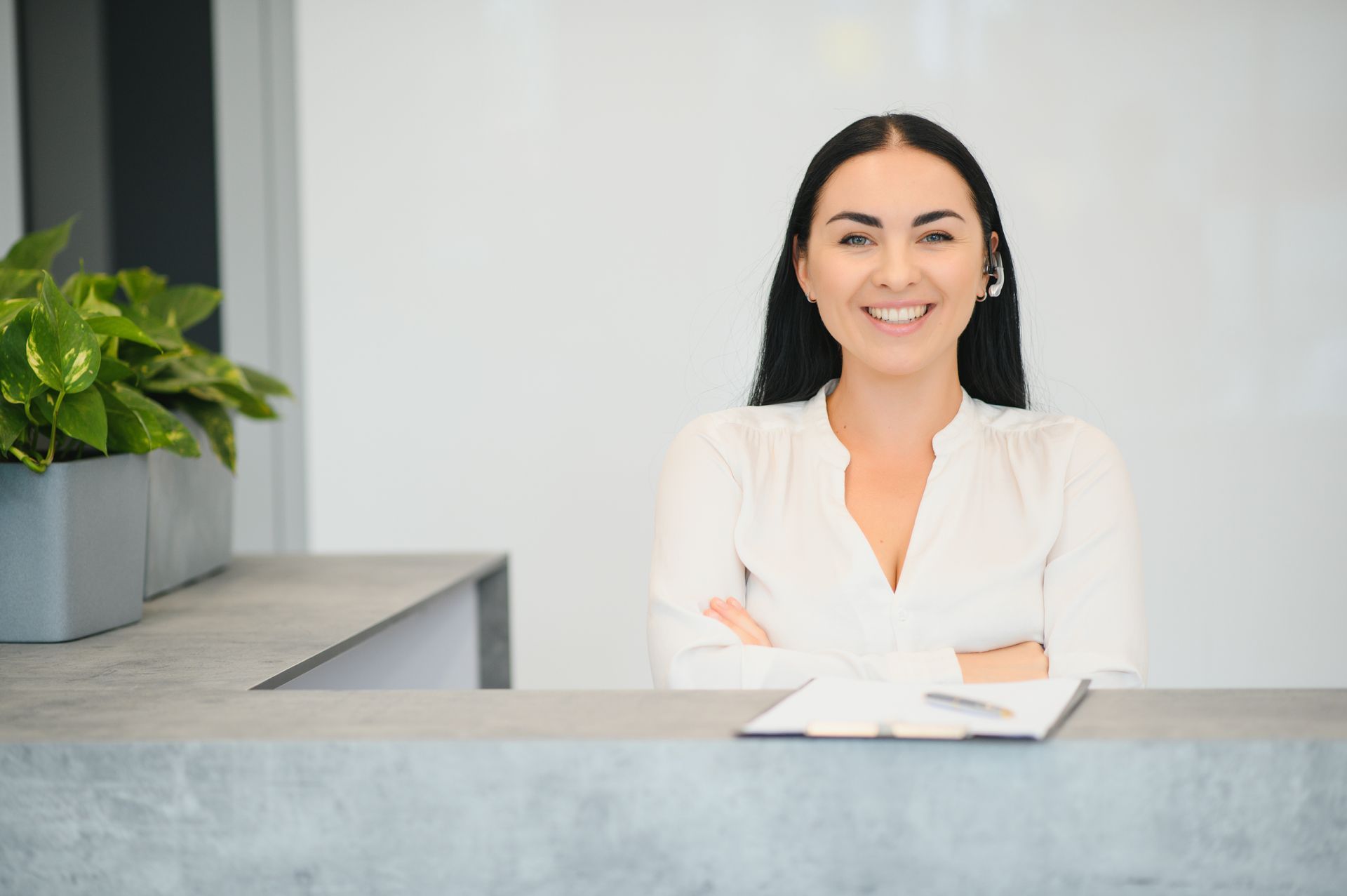 A woman is sitting at a reception desk with her arms crossed and smiling.