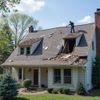 Man on damaged roof of a house, debris scattered. Blue sky, brick chimney, white siding.