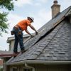 Roofer in orange shirt and hard hat repairs damaged asphalt shingle roof.