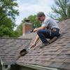 Roofer inspecting shingle roof, holding a tool. Man wearing cap and safety harness.