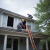 Two construction workers on a roof, one on a ladder, working on a house with a blue exterior.