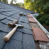 Hammer on a shingle roof near a gutter, with trees in the background.