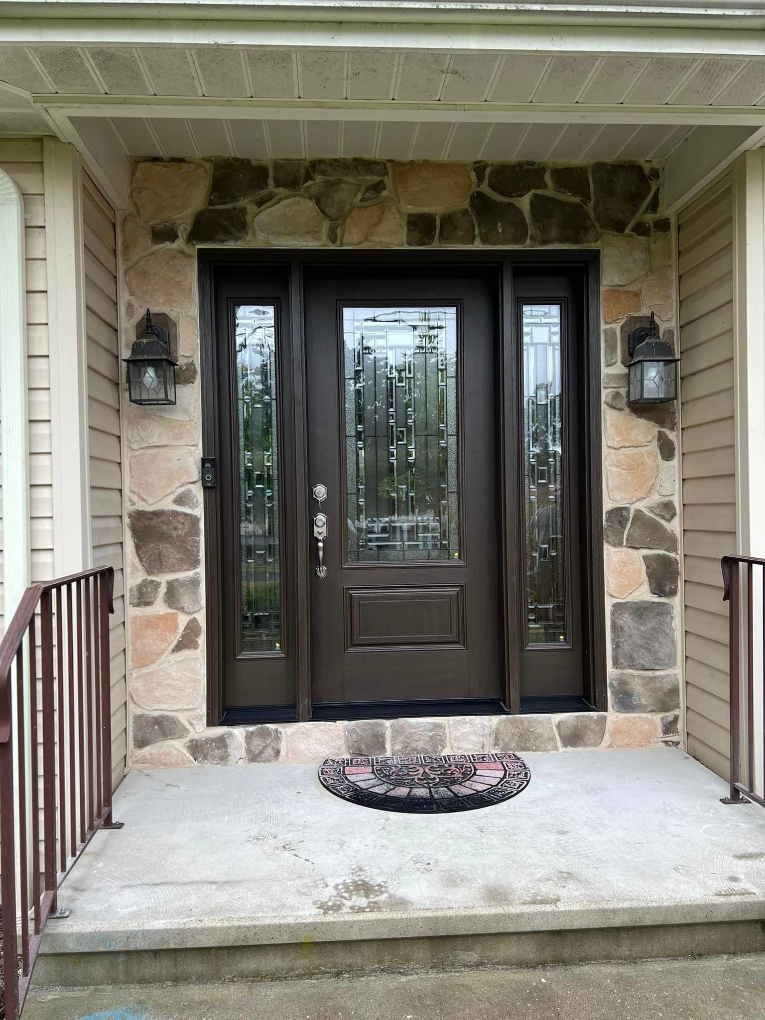 A dark brown front door with glass panels and sidelights, framed by stone, under a porch roof. Two black sconces flank the door.