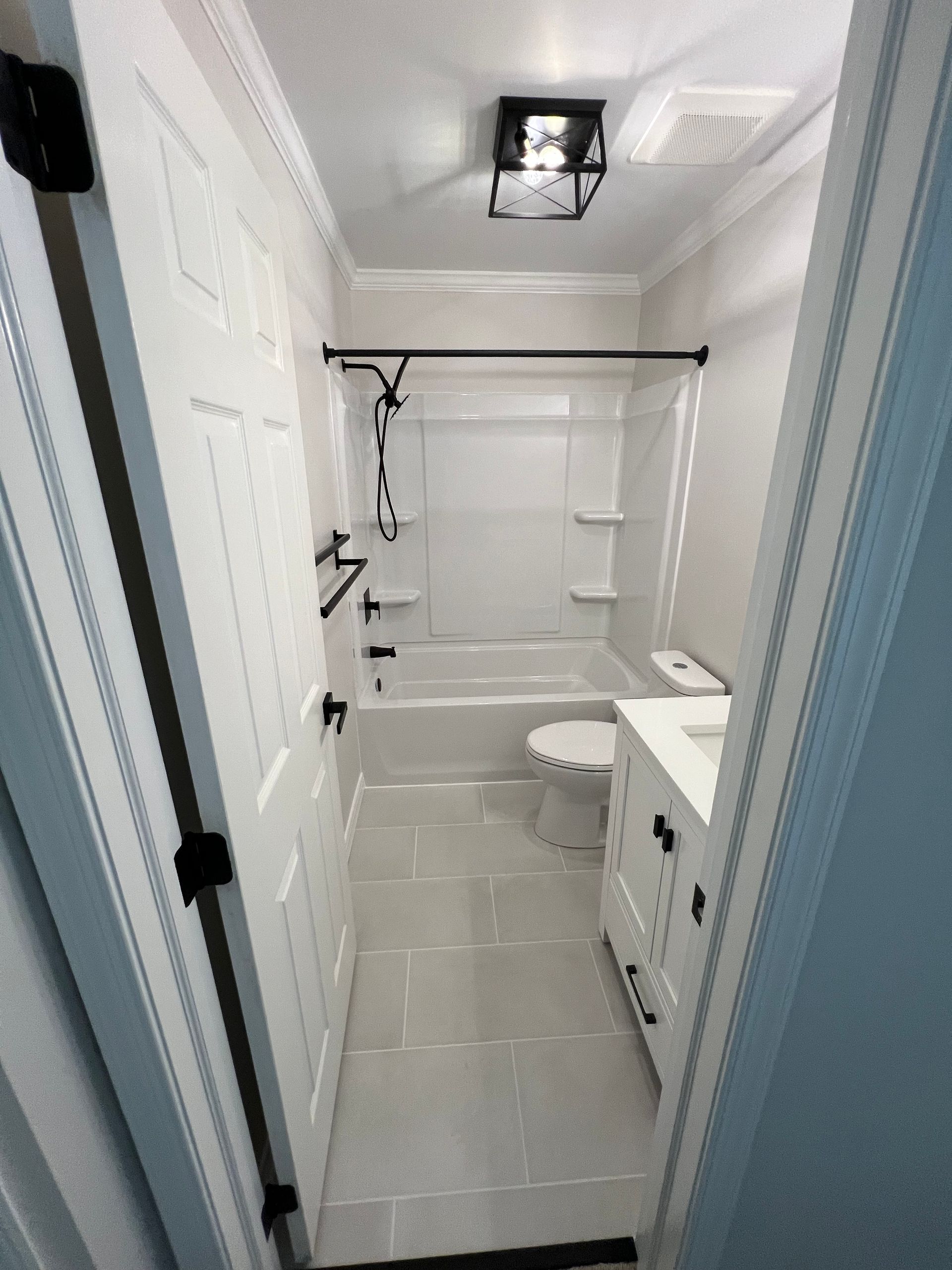 Narrow bathroom with white fixtures, including a tub/shower, toilet, and vanity. Black hardware contrasts with the white walls and tile floor.