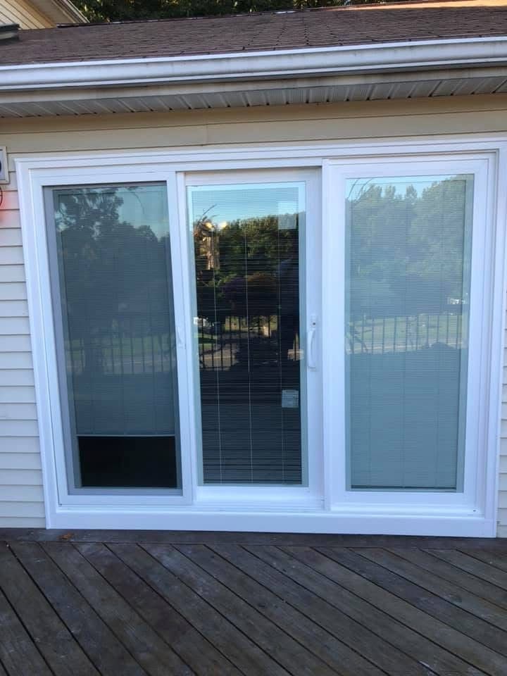 White sliding glass door with two stationary panels and a central door, on a wooden deck. Exterior shot with a light-colored house siding.