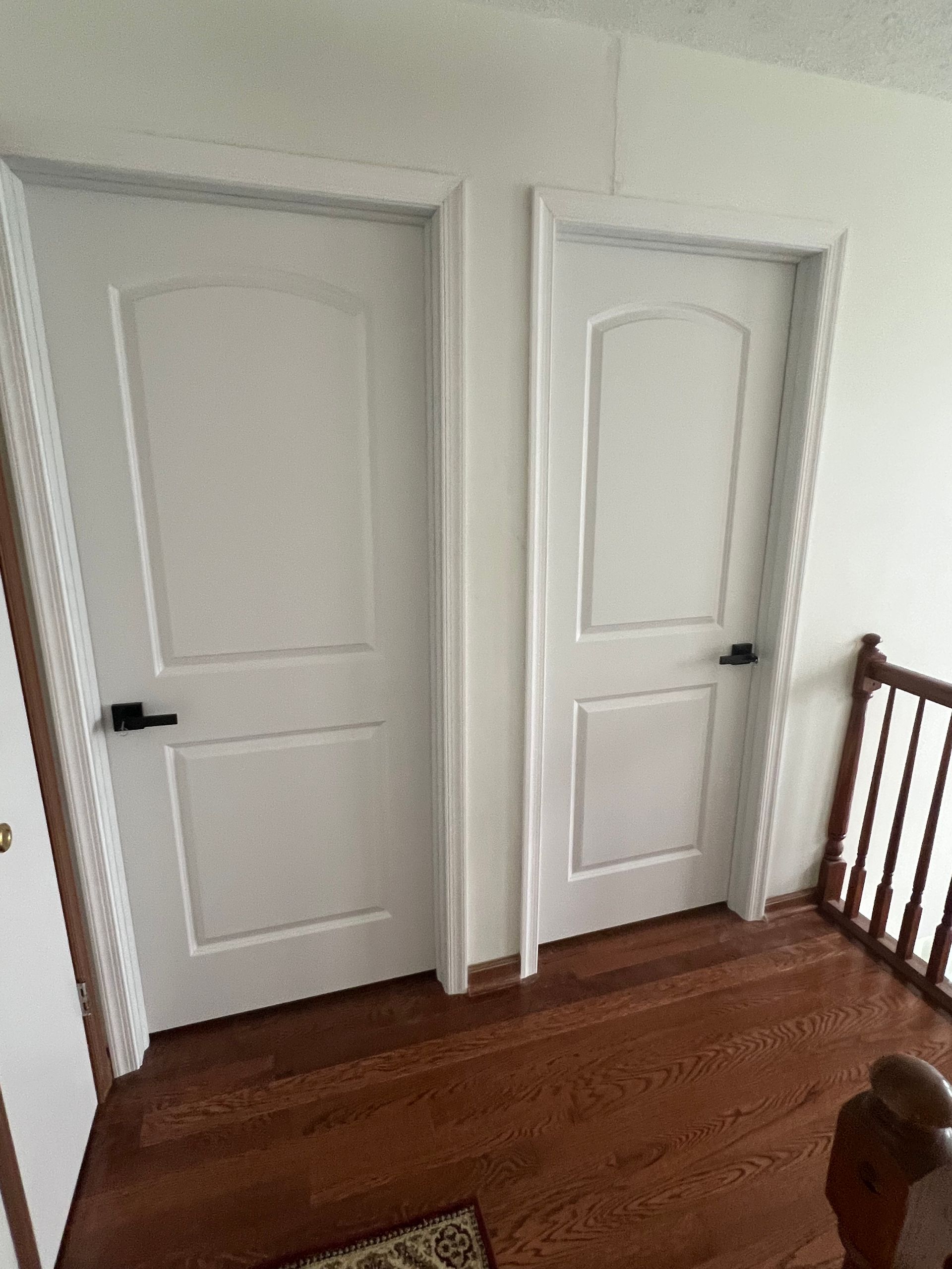 Two white interior doors with black handles stand side-by-side on a hardwood floor near a staircase.