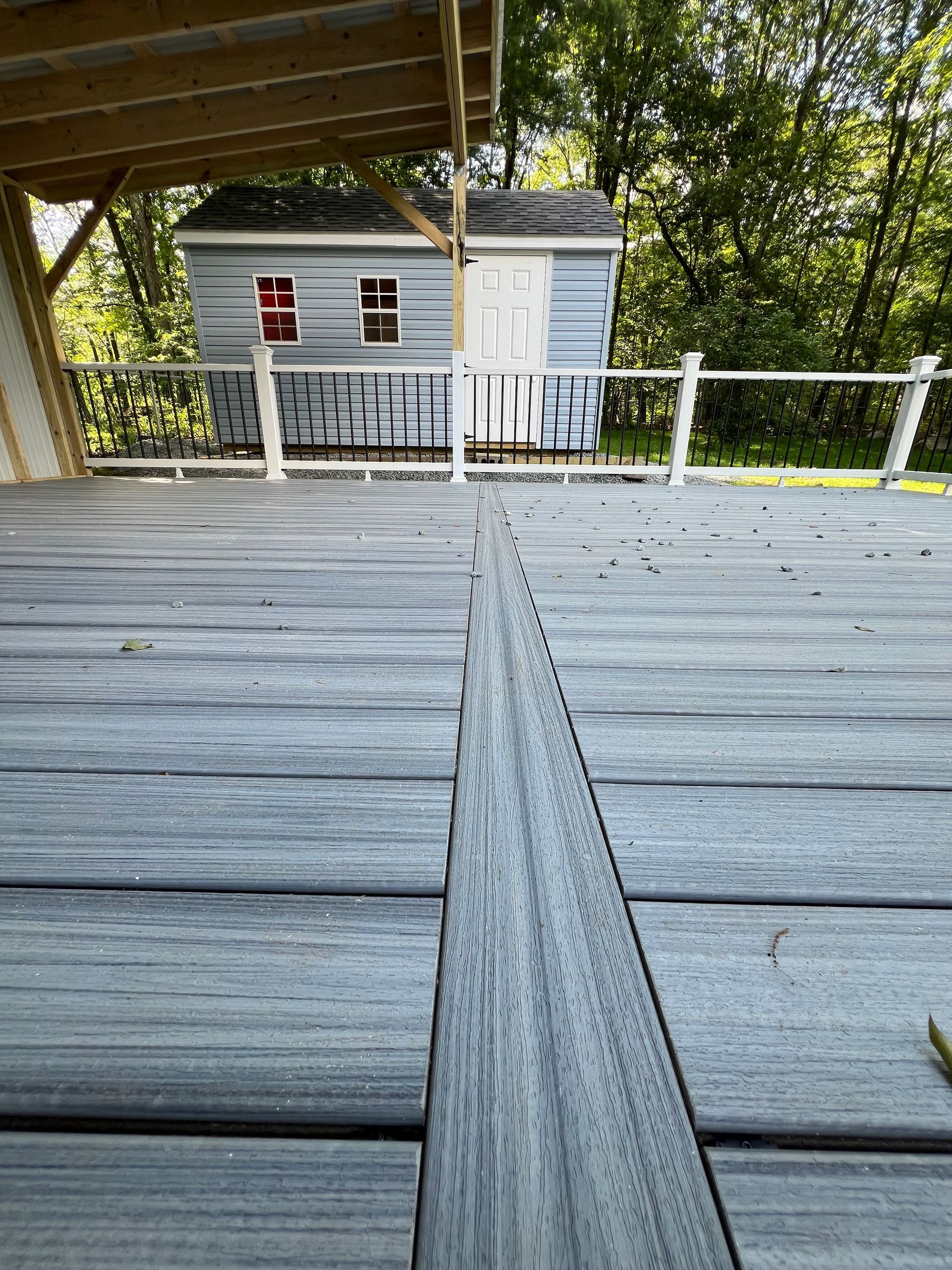 Gray composite deck leads to a light blue shed with white trim, set in a wooded area. A covered porch is on the left.