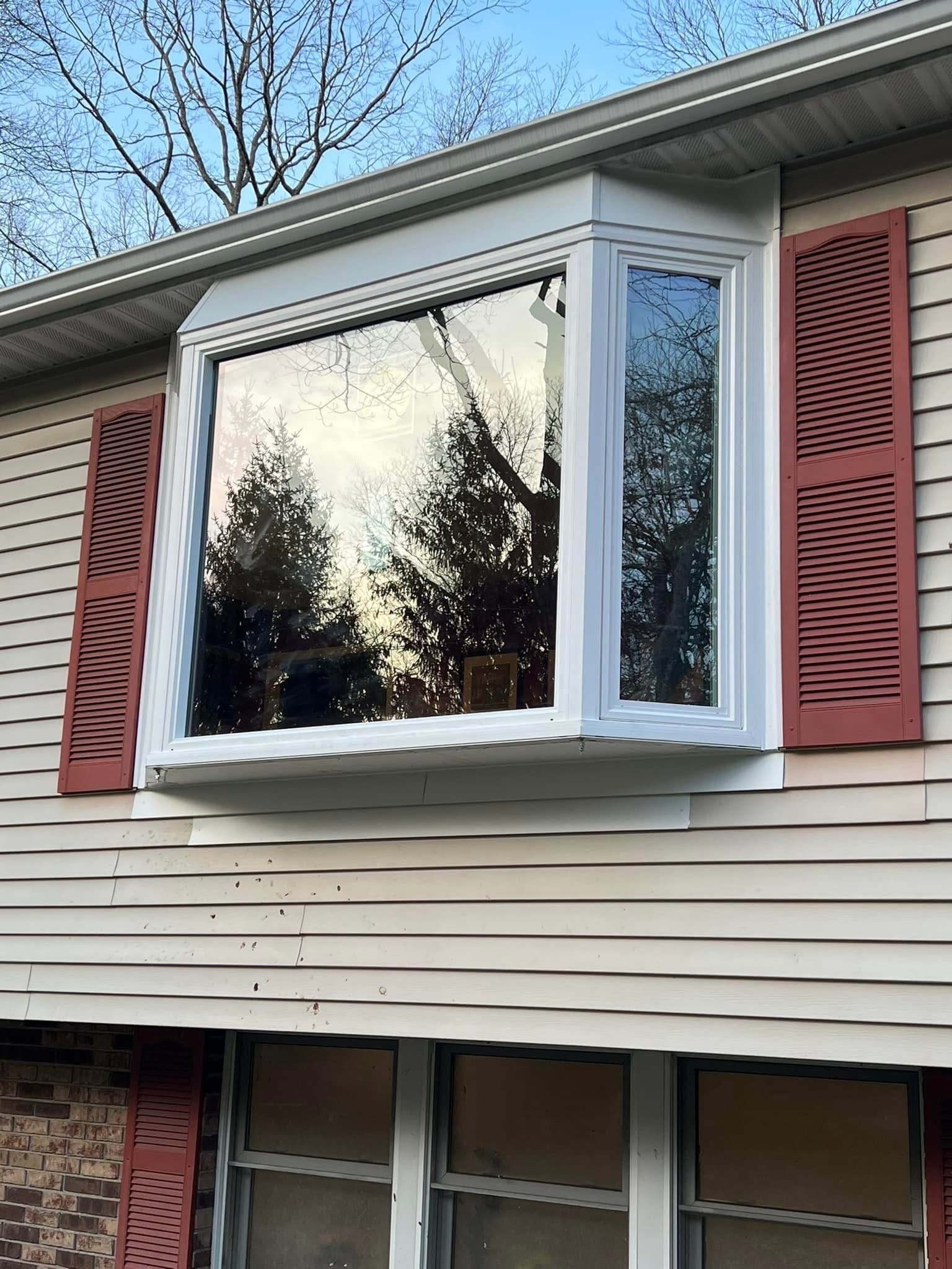 A bay window on a beige house reflects trees and sky, with reddish-brown shutters on either side.