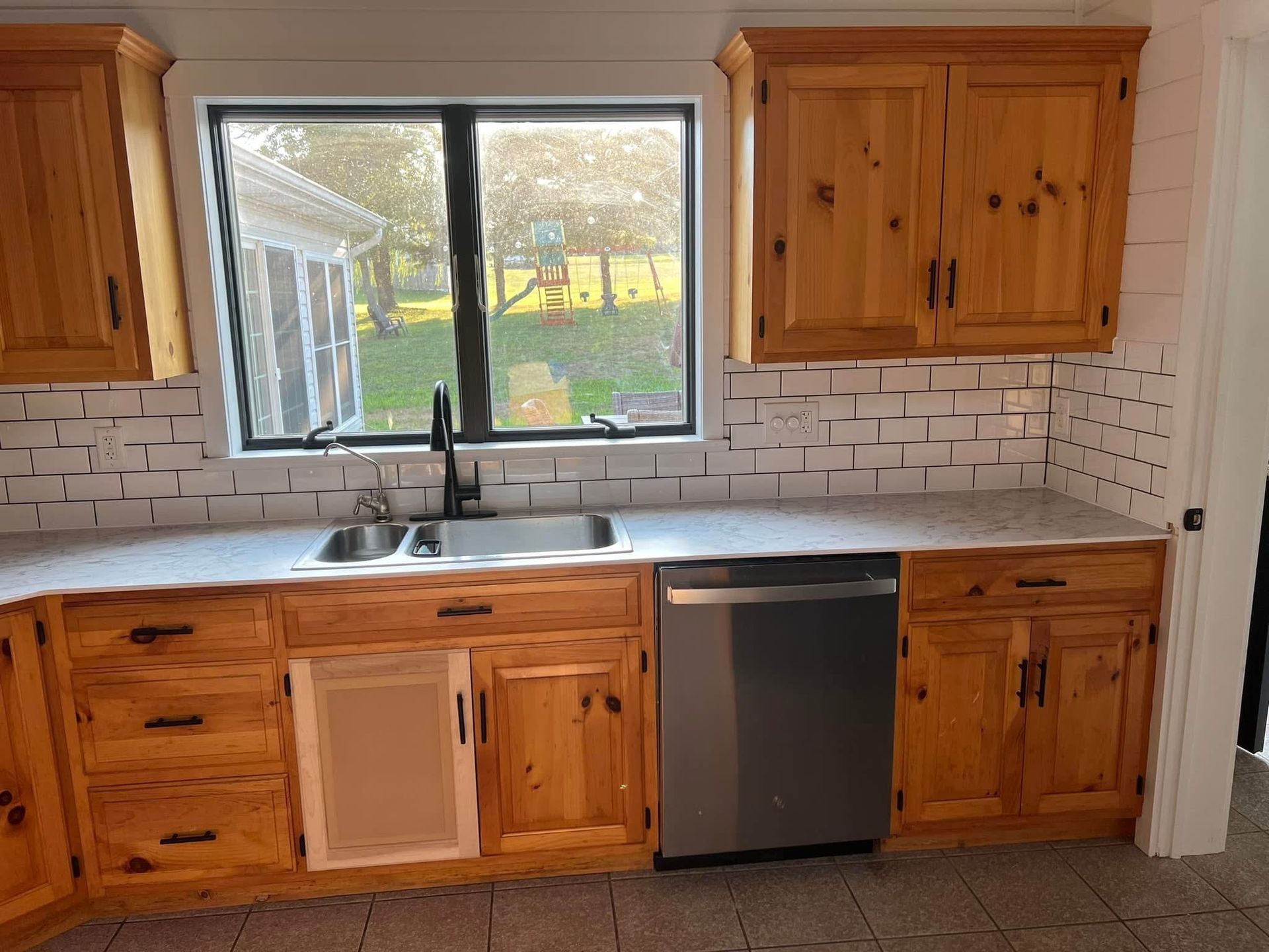 Kitchen with light wood cabinets, a stainless steel dishwasher, and a black sink and faucet, with a window looking out to a backyard.