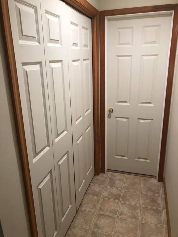 Narrow hallway with a white bi-fold closet door on the left and a white door on the right with brown trim. Beige tiled floor.