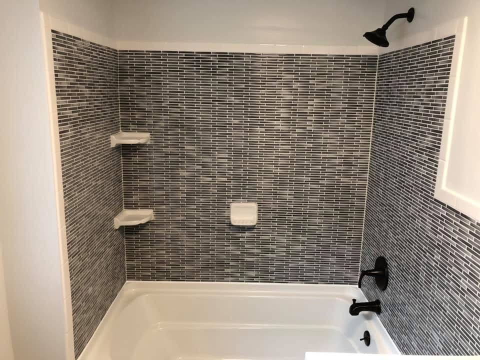 A small bathroom with a white tub and patterned black and white tiled shower walls, black fixtures, and white shelves.