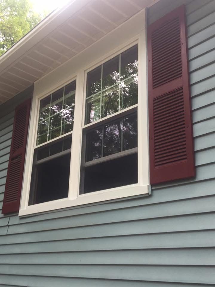 A window with white trim and red shutters on a blue-sided house, reflecting trees.
