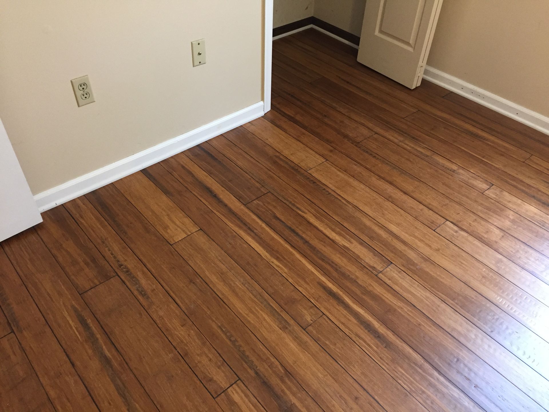 Wooden floor in a room with beige walls and white trim. An electrical outlet is visible on the wall.