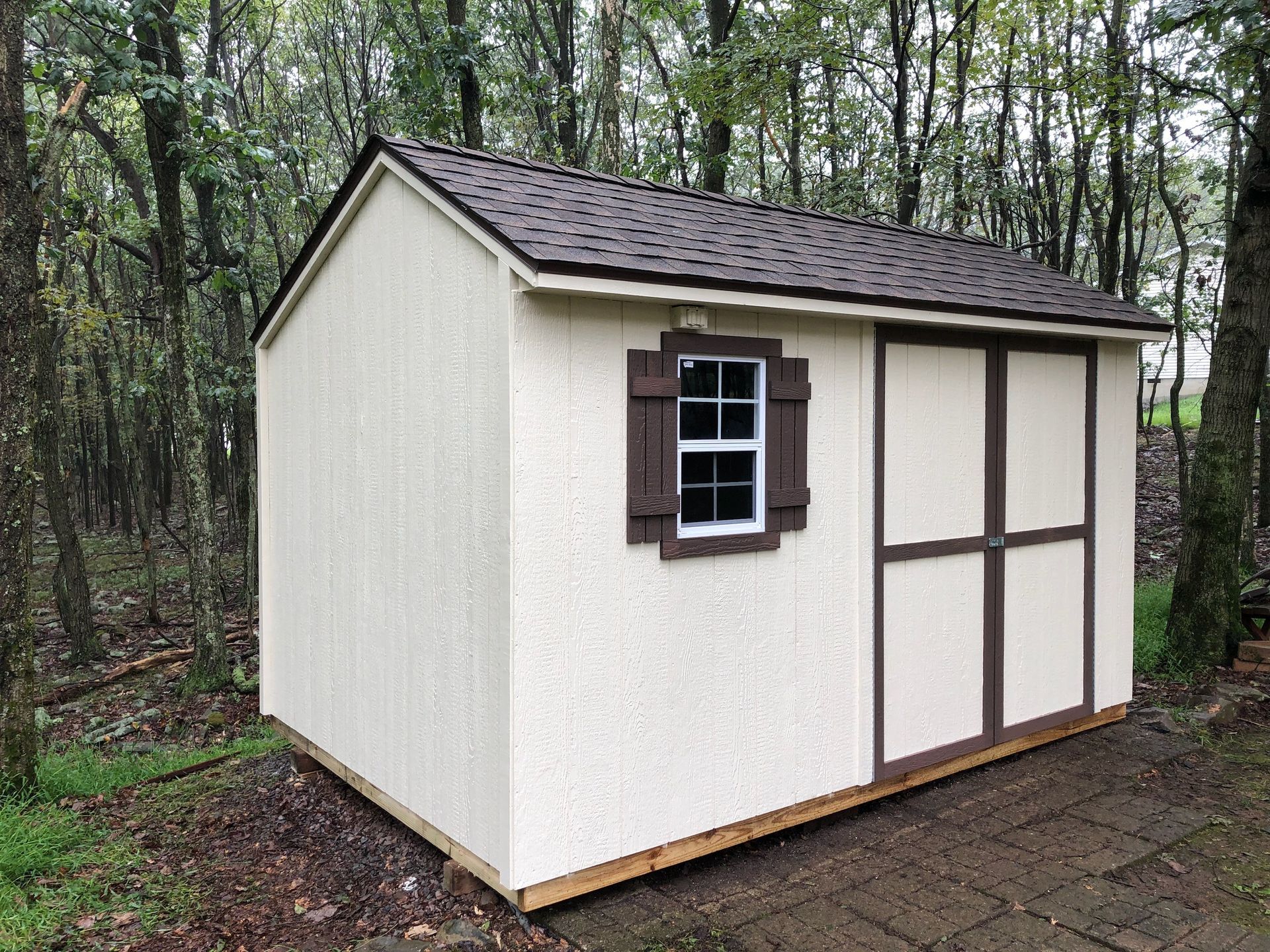 A tan and brown shed with a window and sliding door stands in a wooded area.