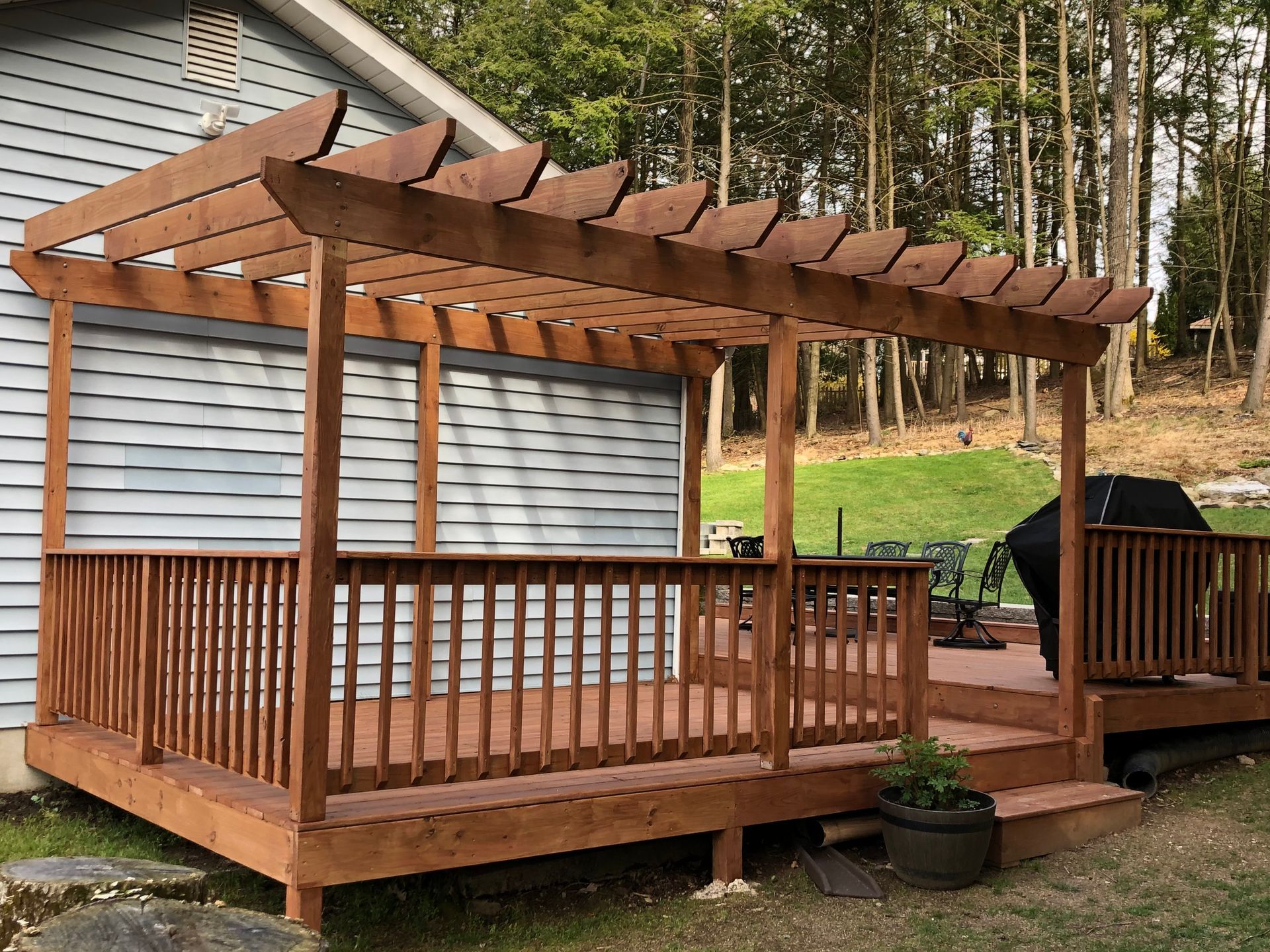 Wooden deck and pergola attached to a light blue house, with a grassy yard and trees in the background. The deck has a railing and stairs.