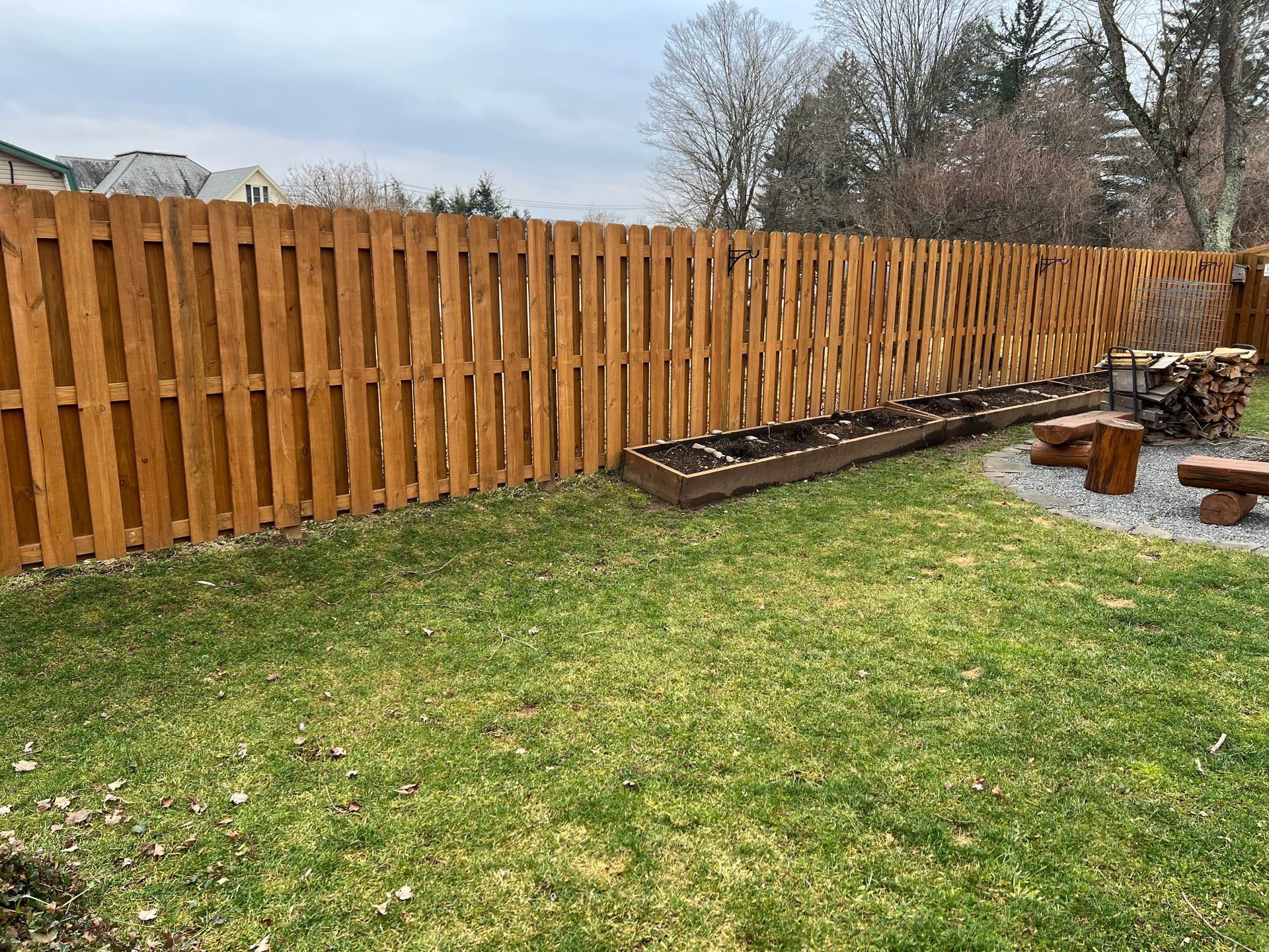 Wooden fence surrounding a backyard with patchy green grass. A small garden bed and log benches are visible.