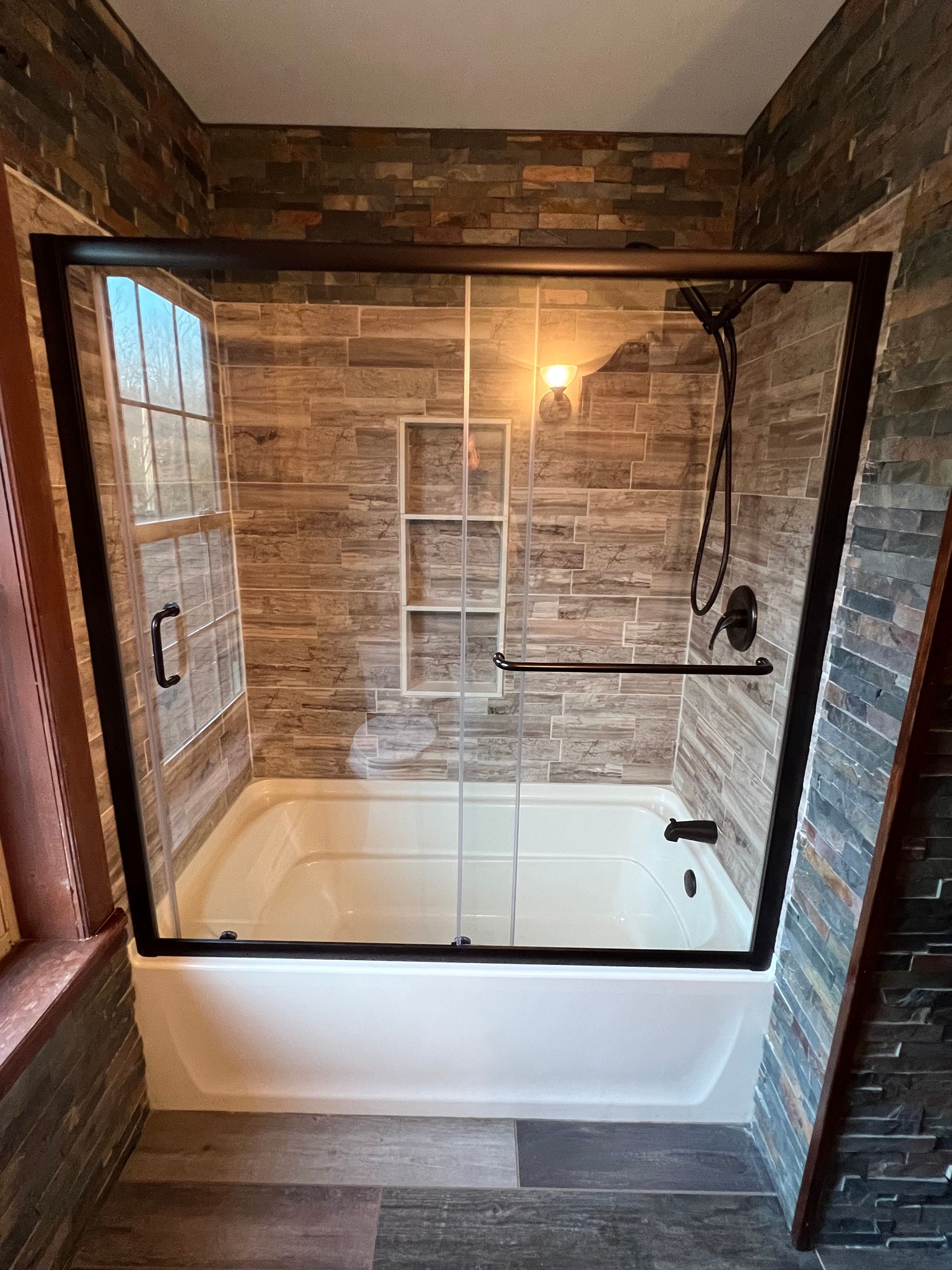 Bathroom with a white tub, dark framed shower door, and stone tile walls.