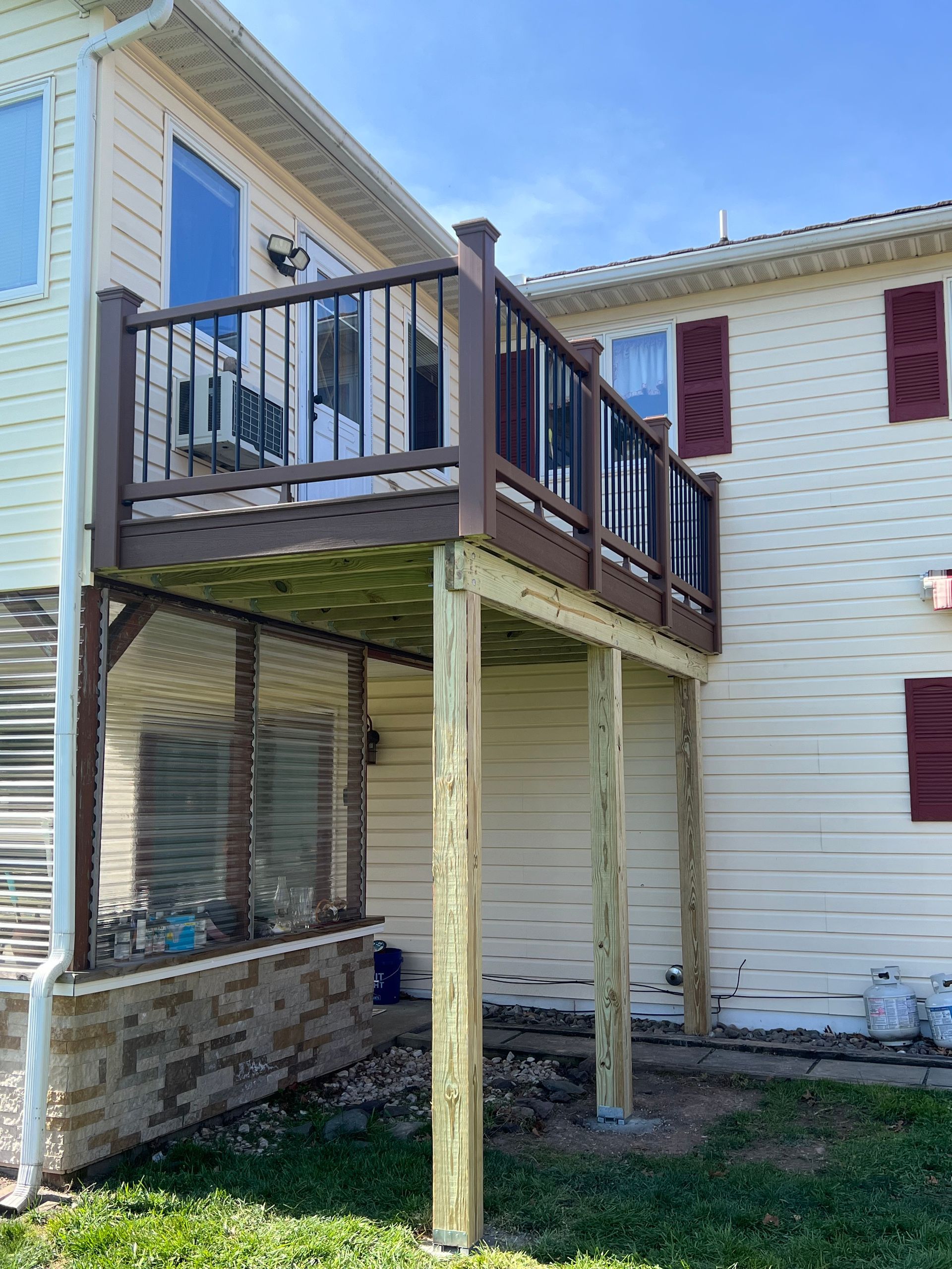 A raised wooden deck attached to a two-story house. The deck has brown railings and supports, overlooking a lower area with stone siding.