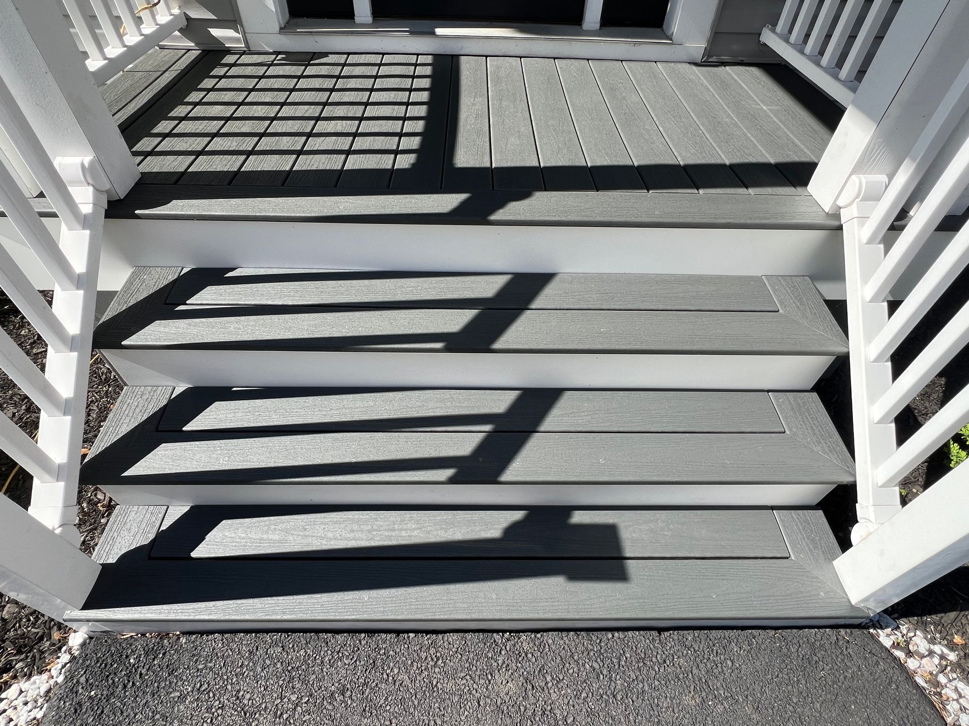Gray composite steps leading up to a doorway, with white railings on either side. Sunlight casts shadows.