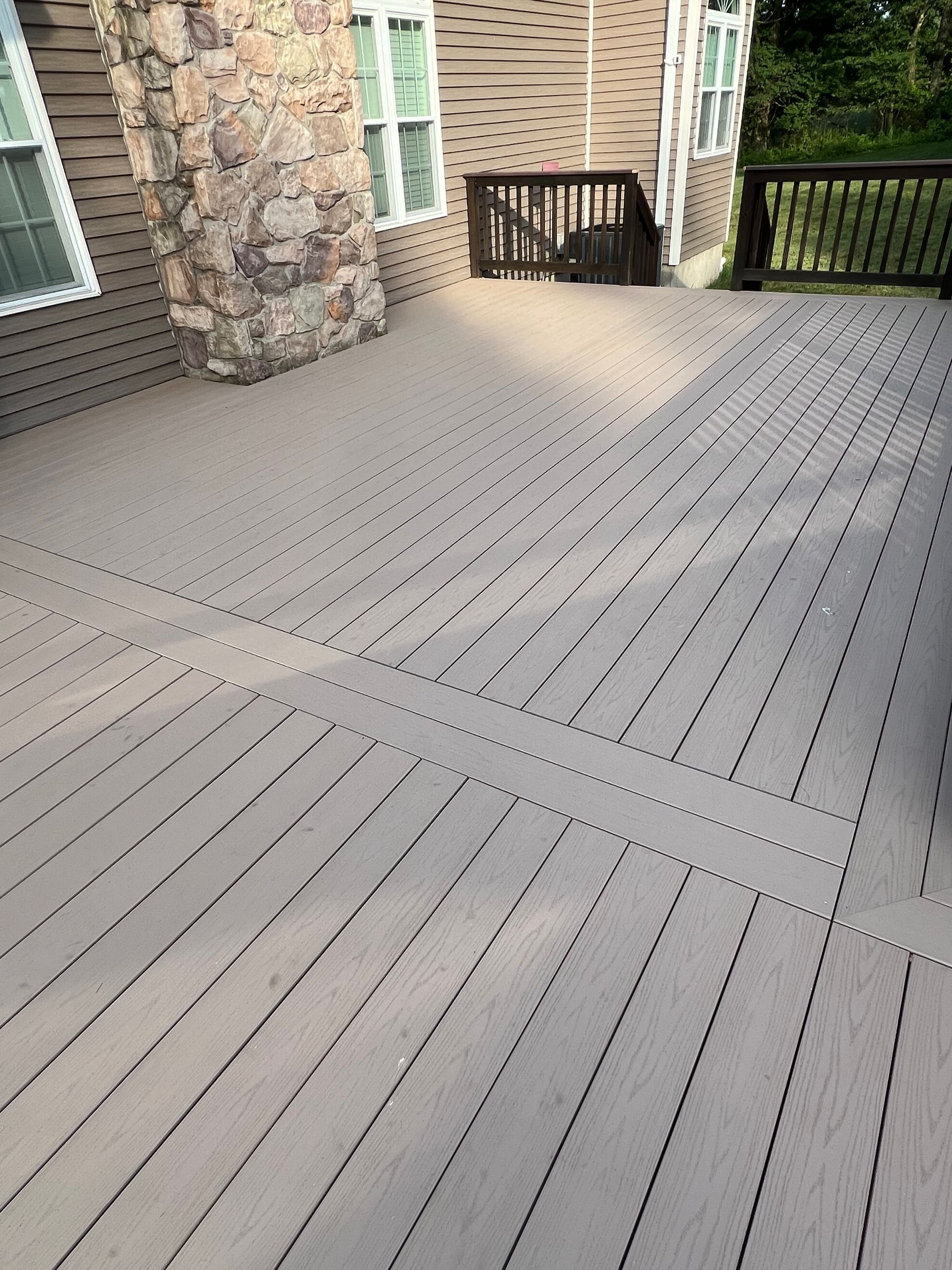 A weathered wooden deck with light-brown boards, part of a house with stone accents and windows. The deck leads to a small shaded area.