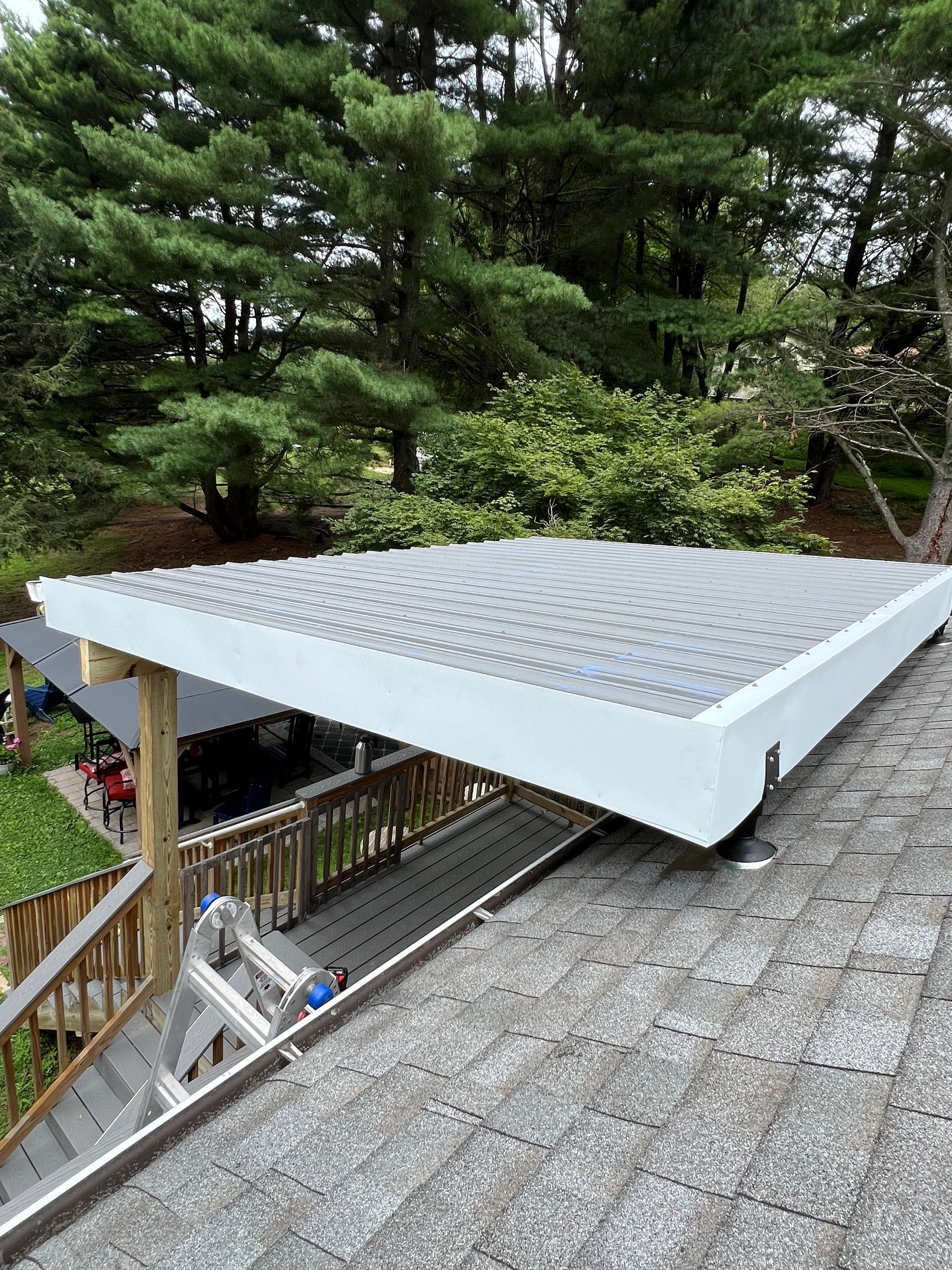 White-trimmed carport with a gray, patterned roof extending over a wooden deck, viewed from a rooftop perspective. Trees and a garden are visible in the background.