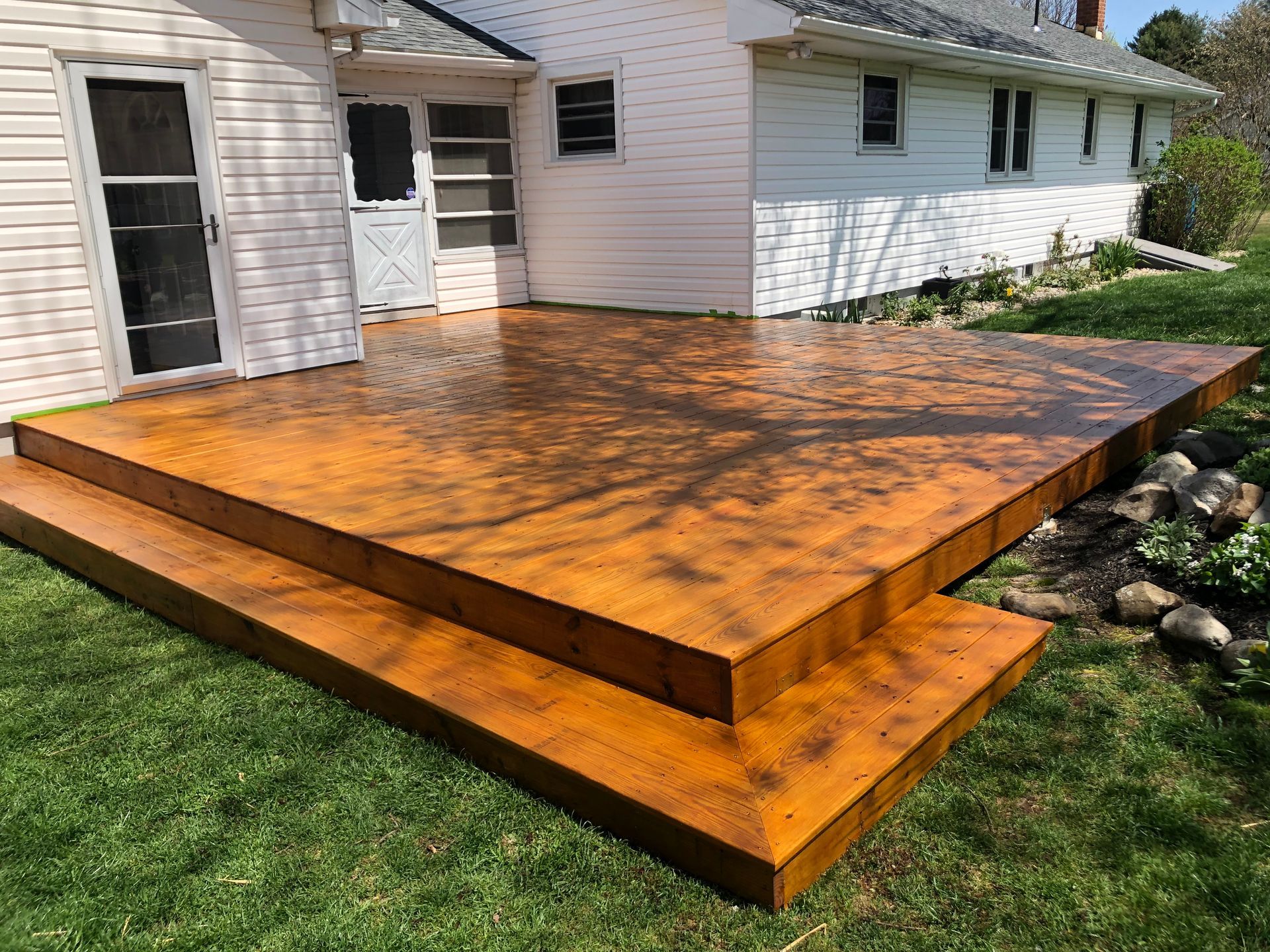 Wooden deck with two steps, freshly stained, next to a white house on a grassy lawn. Tree shadows are cast across the surface.