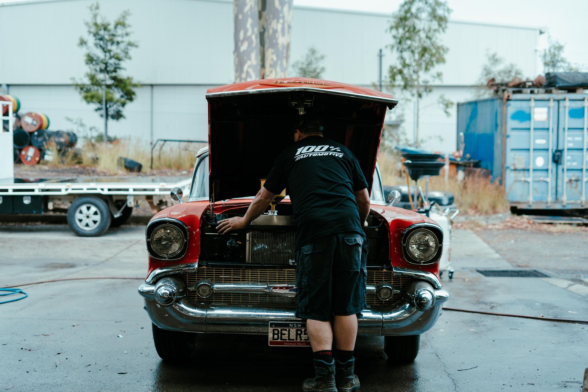 Person in black shirt working on a red classic car with open hood, in a wet outdoor setting. β 100% Automotive In South Nowra, NSW