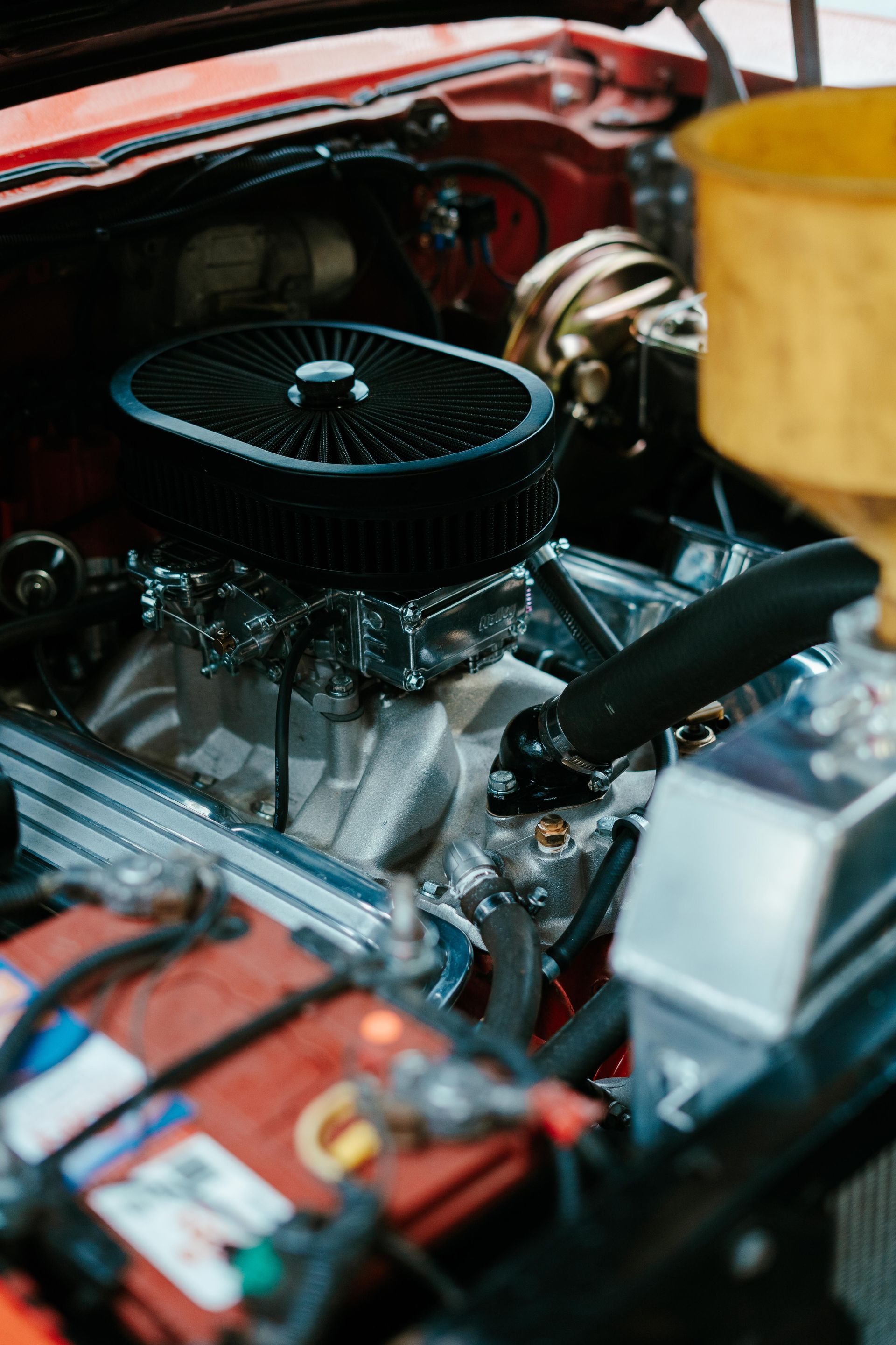 Close-up of a classic car engine; black air filter on a silver engine block, red battery. — 100% Automotive In South Nowra, NSW