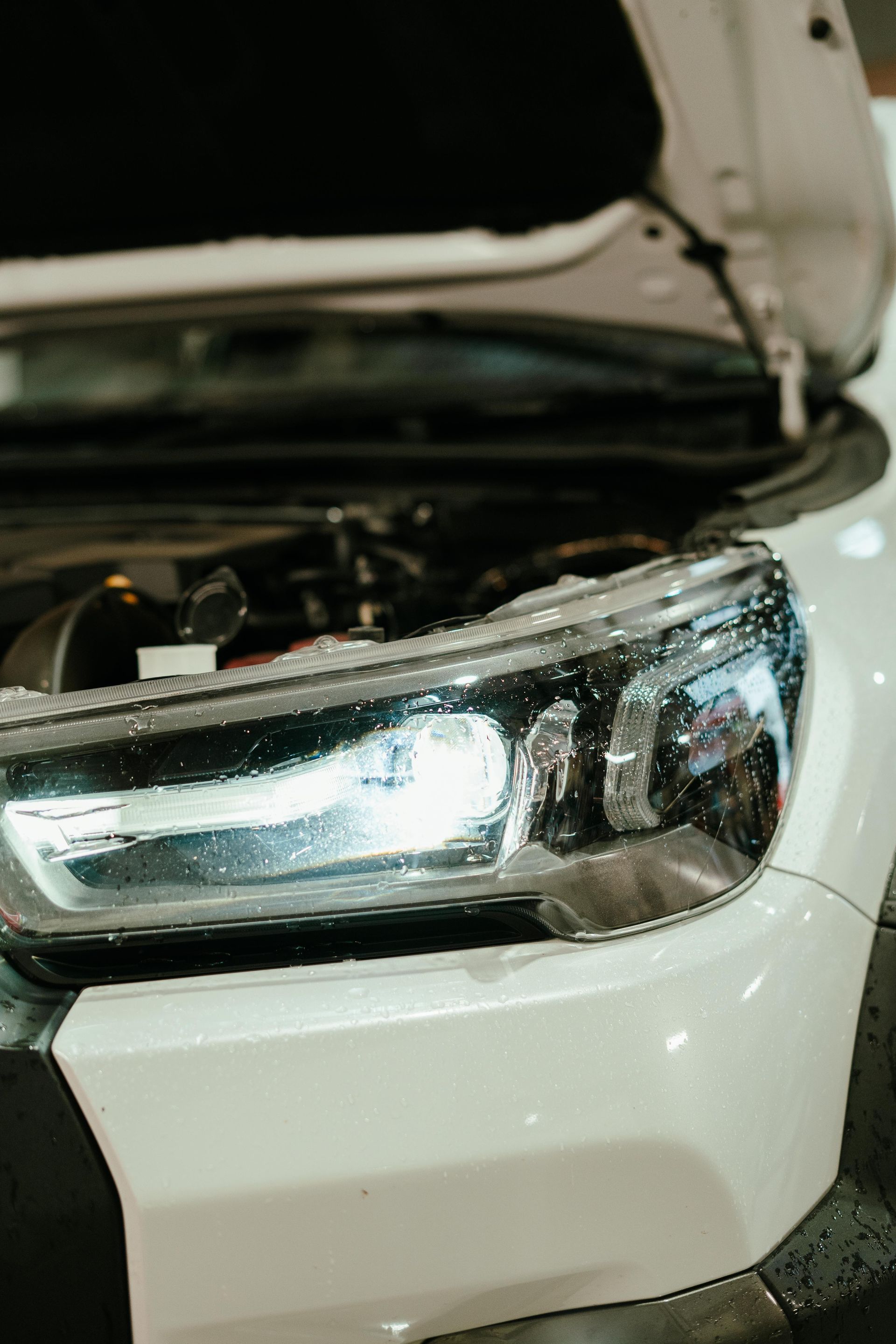 Close-up of a white car's headlight illuminated with the hood open, revealing the engine. — 100% Automotive In South Nowra, NSW