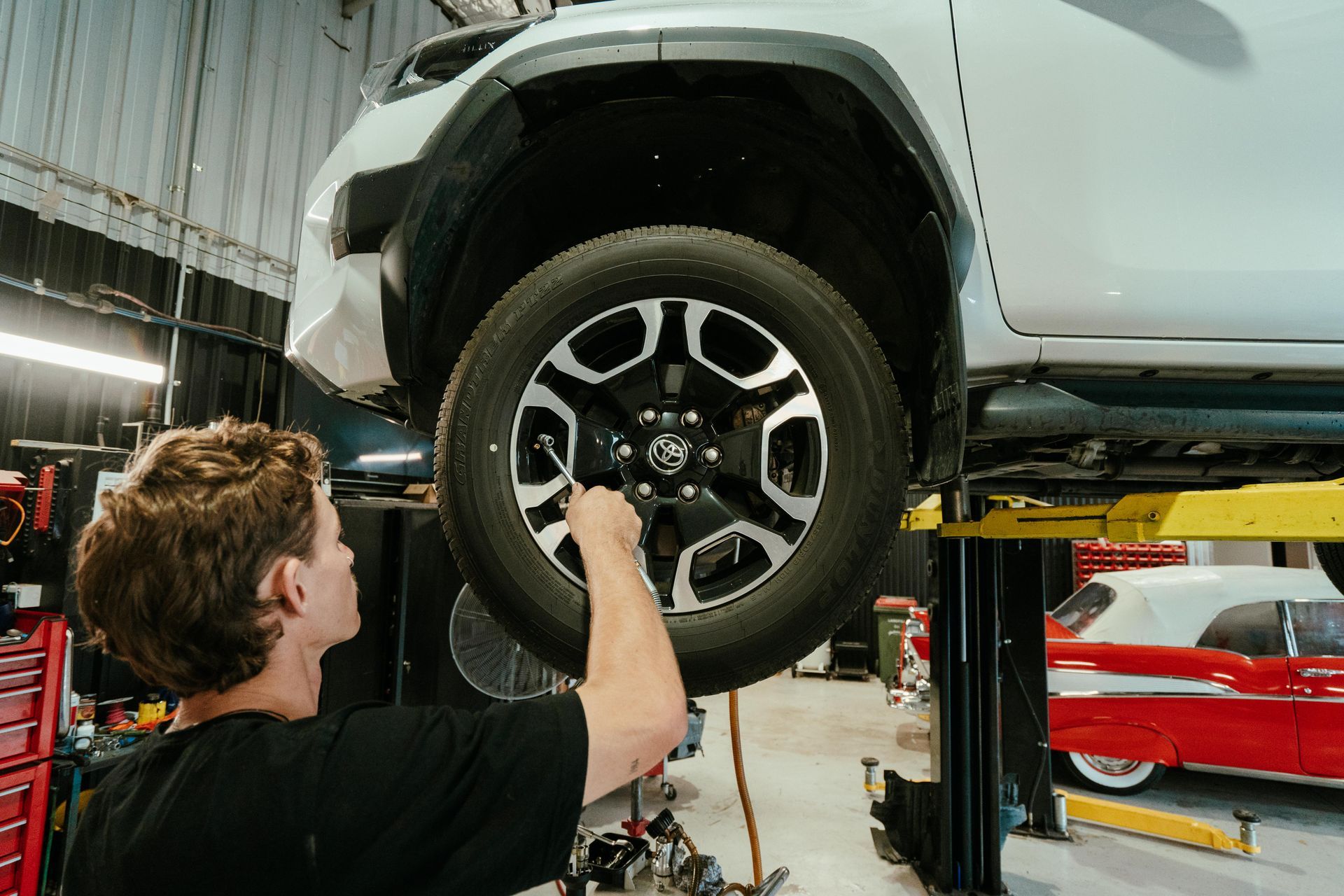Mechanic working on the tire of a white truck in a garage; red toolbox and classic car visible. β 100% Automotive In South Nowra, NSW