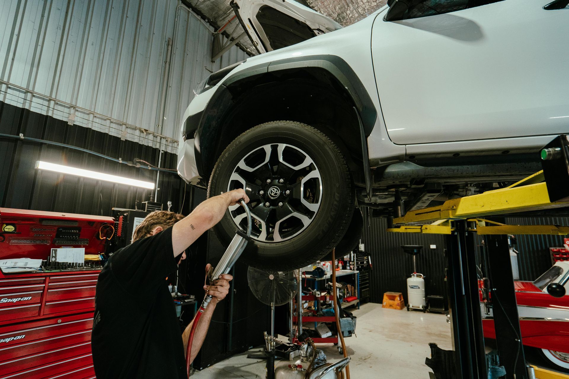 Mechanic working on a white vehicle raised on a lift inside a garage, using a tool on the wheel. — 100% Automotive In South Nowra, NSW