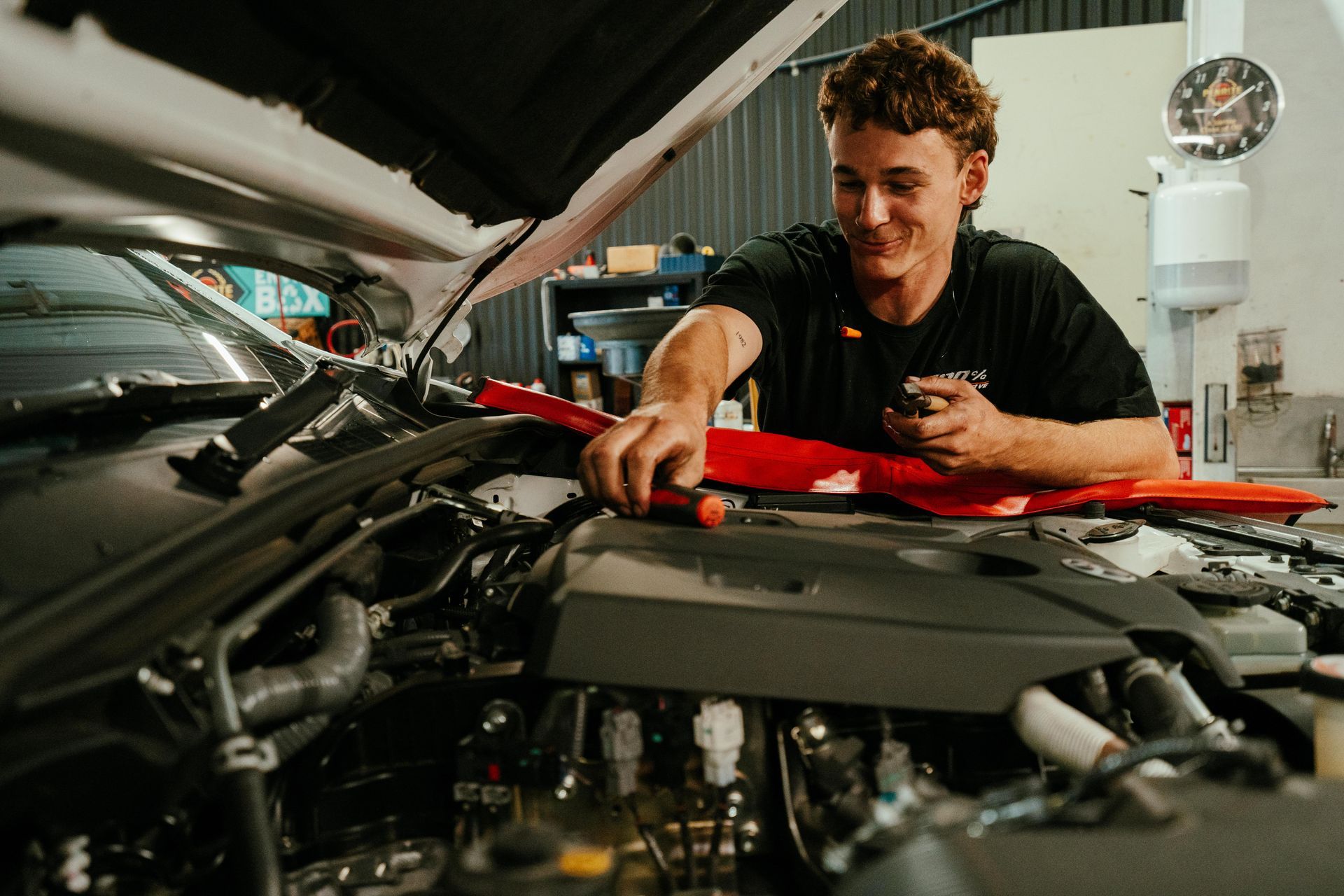 Mechanic smiles, working on a car engine under the hood in a garage, using a red tool. β 100% Automotive In South Nowra, NSW