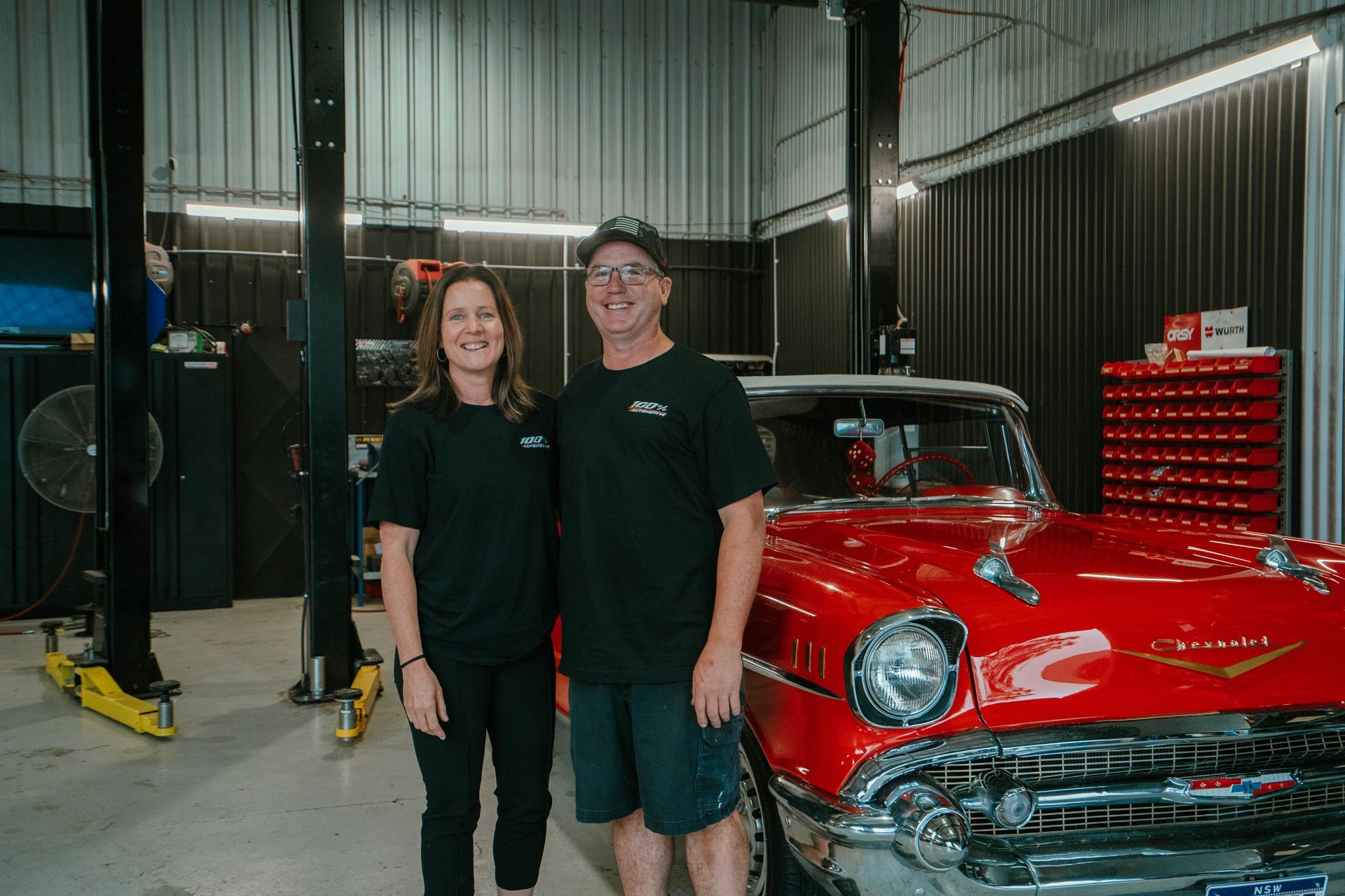 Woman and man stand beside a bright red classic car in an auto shop. — 100% Automotive In South Nowra, NSW