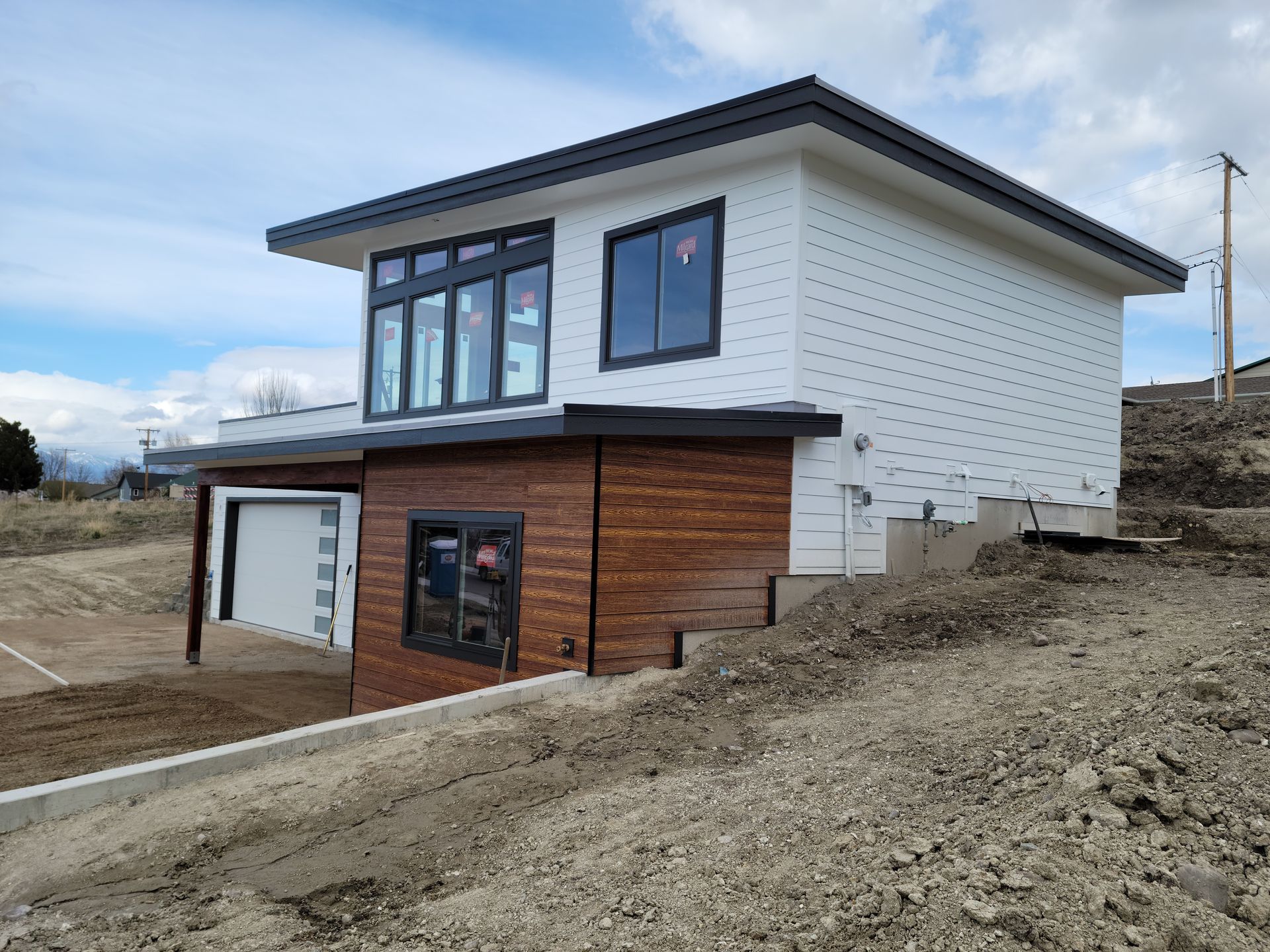 Modern two-story house with white siding, wood accents, and a garage on a dirt lot under a cloudy sky