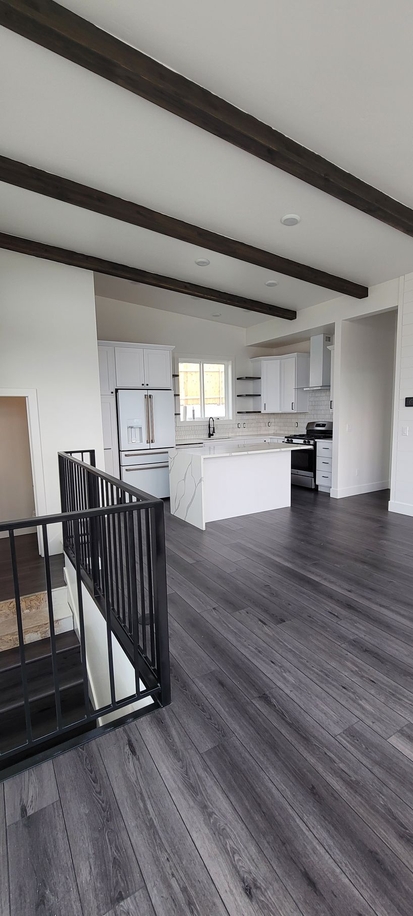 Modern open-plan interior with dark wood beams, gray floors, white walls, and a kitchen island near stairs.