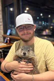 A person wearing a white baseball cap and glasses holds a woodchuck in their arms while sitting in a restaurant.