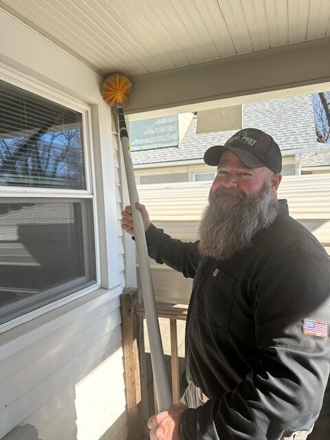 A smiling person with a full beard, wearing a cap, uses an extension pole with a brush to clean a porch ceiling corner.