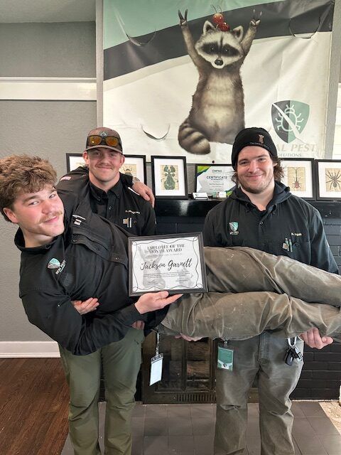 Three staff members in black uniforms stand indoors, holding a framed certificate while one is lifted horizontally.