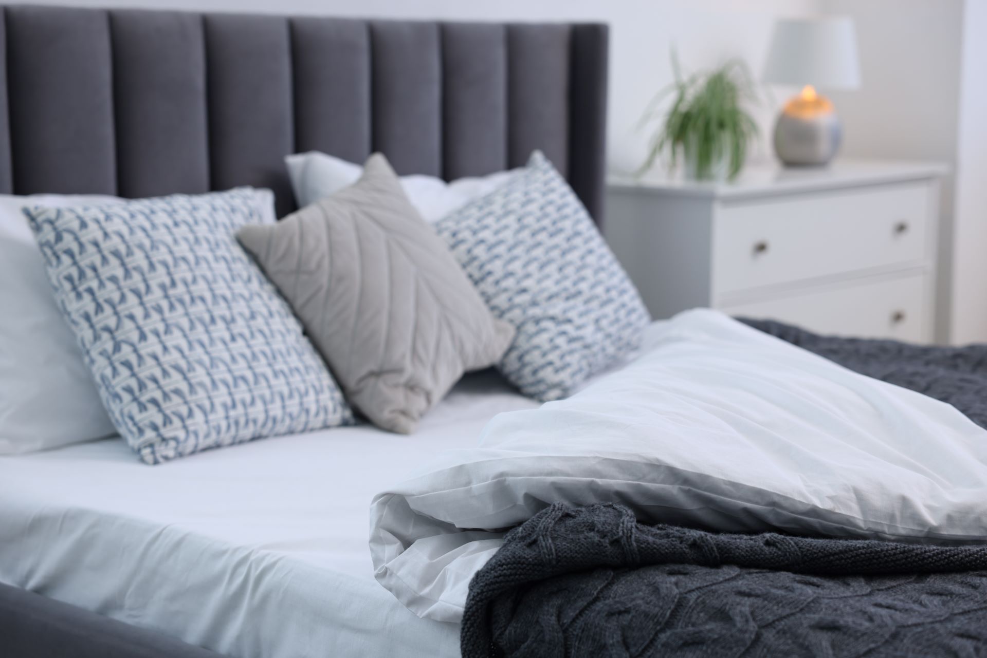 A gray upholstered bed with patterned and solid throw pillows, white bedding, and a dark knit blanket, near a nightstand.