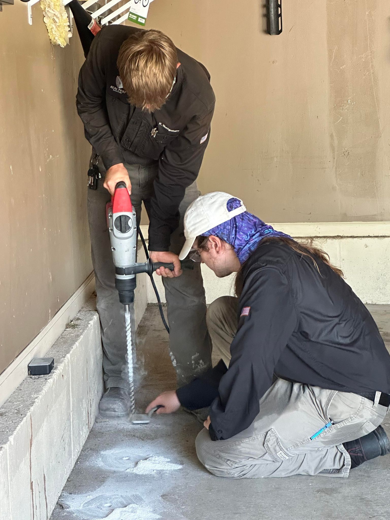 Two people in work clothing use a power drill to bore a hole into a concrete floor alongside a wall.