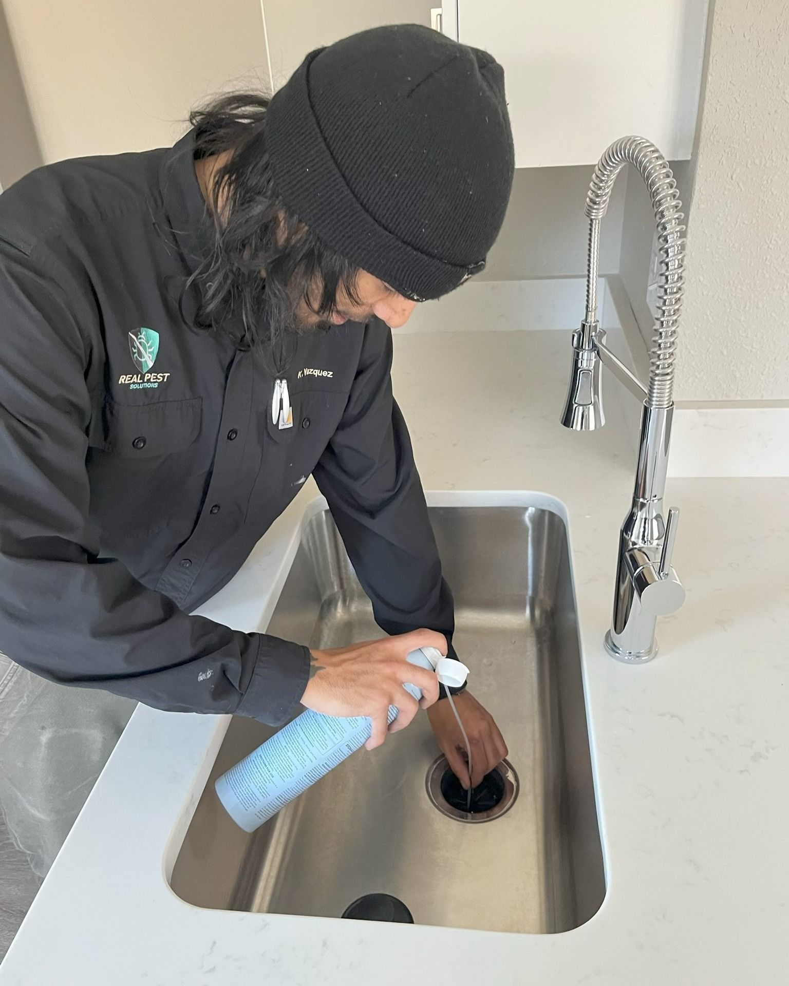 A person in a black shirt and beanie uses a spray cleaner on a kitchen sink drain.