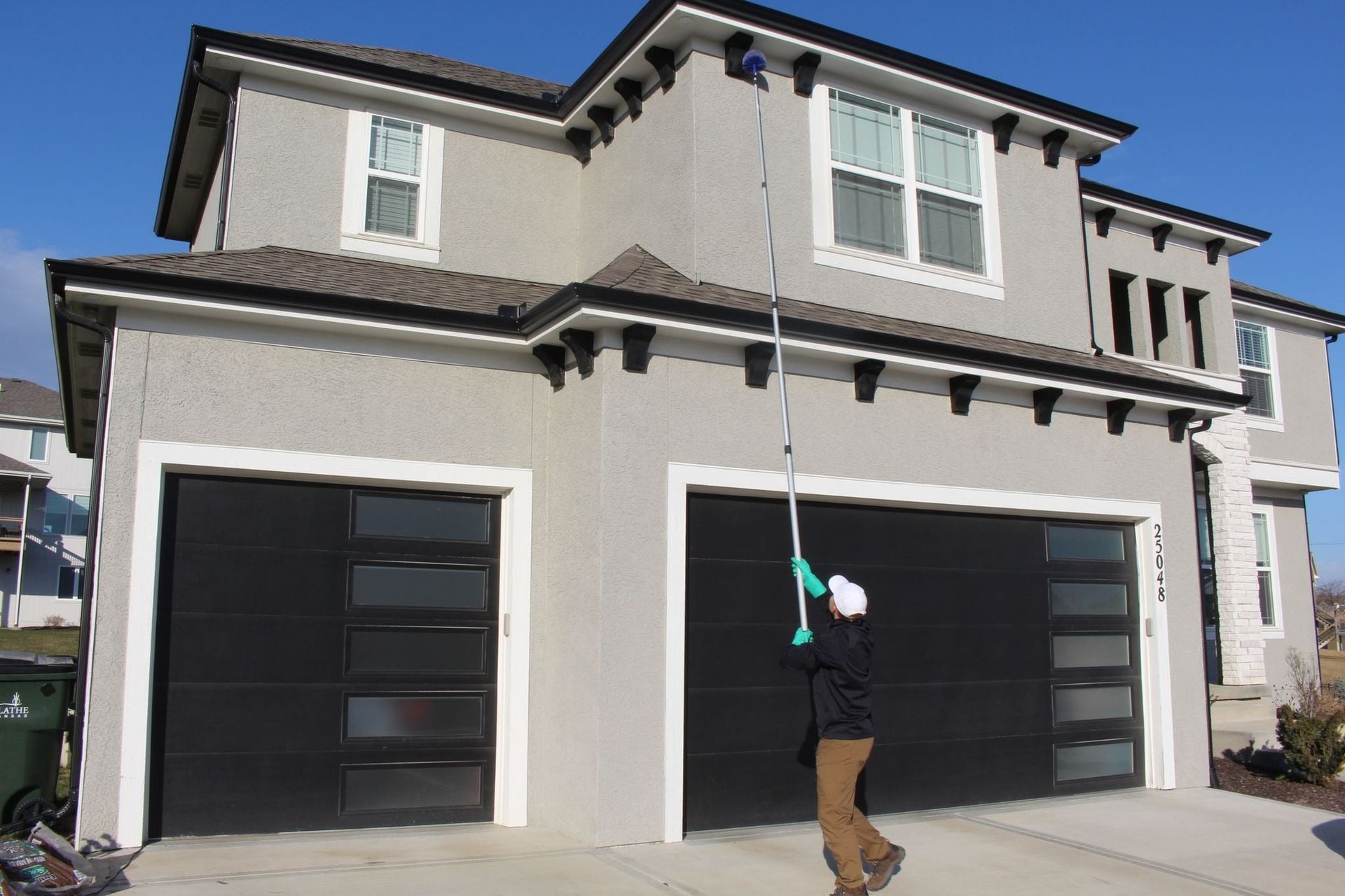 A person stands on a driveway, using a long pole to clean the exterior roofline and trim of a two-story gray house.